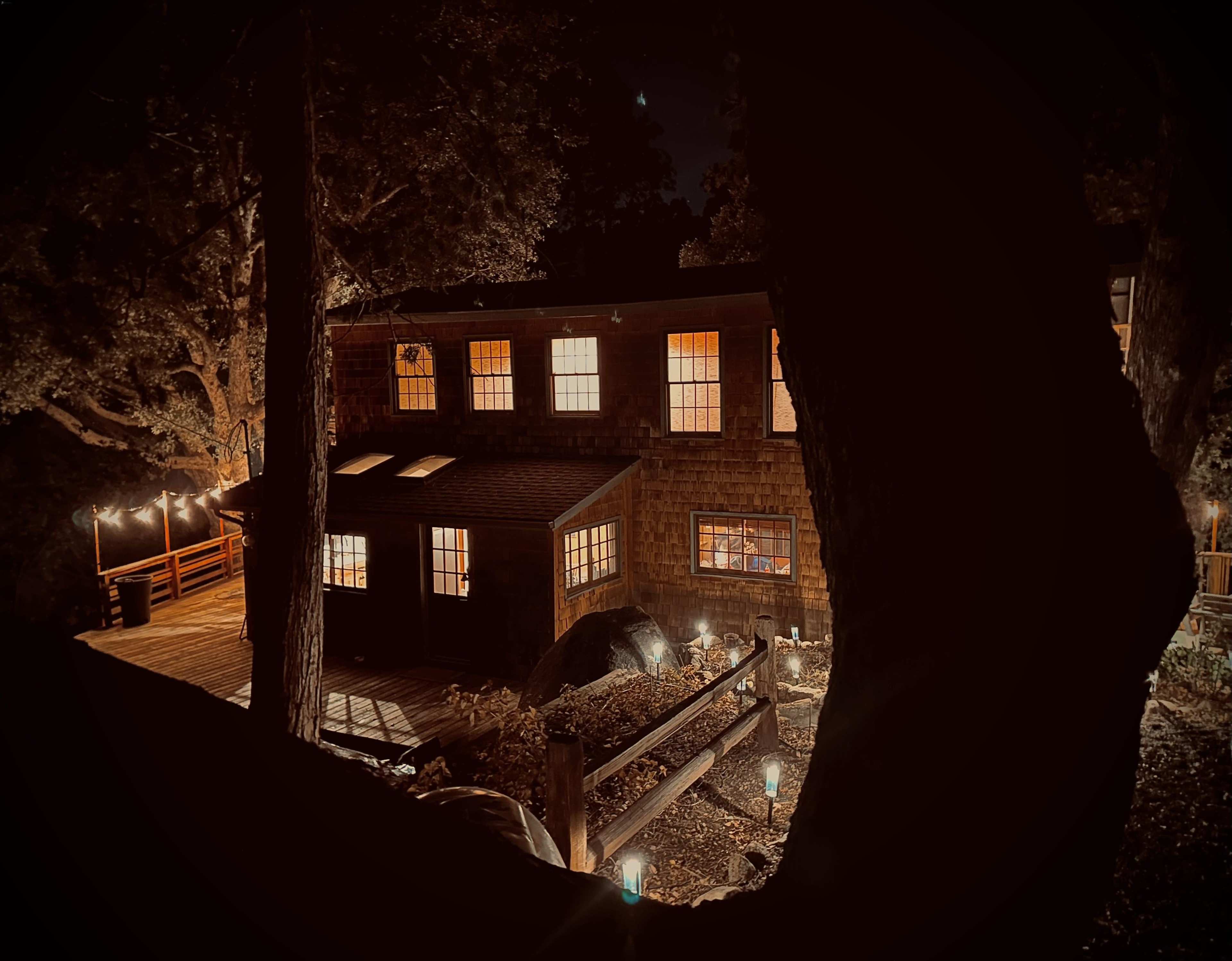 A wooden house with illuminated windows is nestled among trees at night, surrounded by a deck and garden lights.