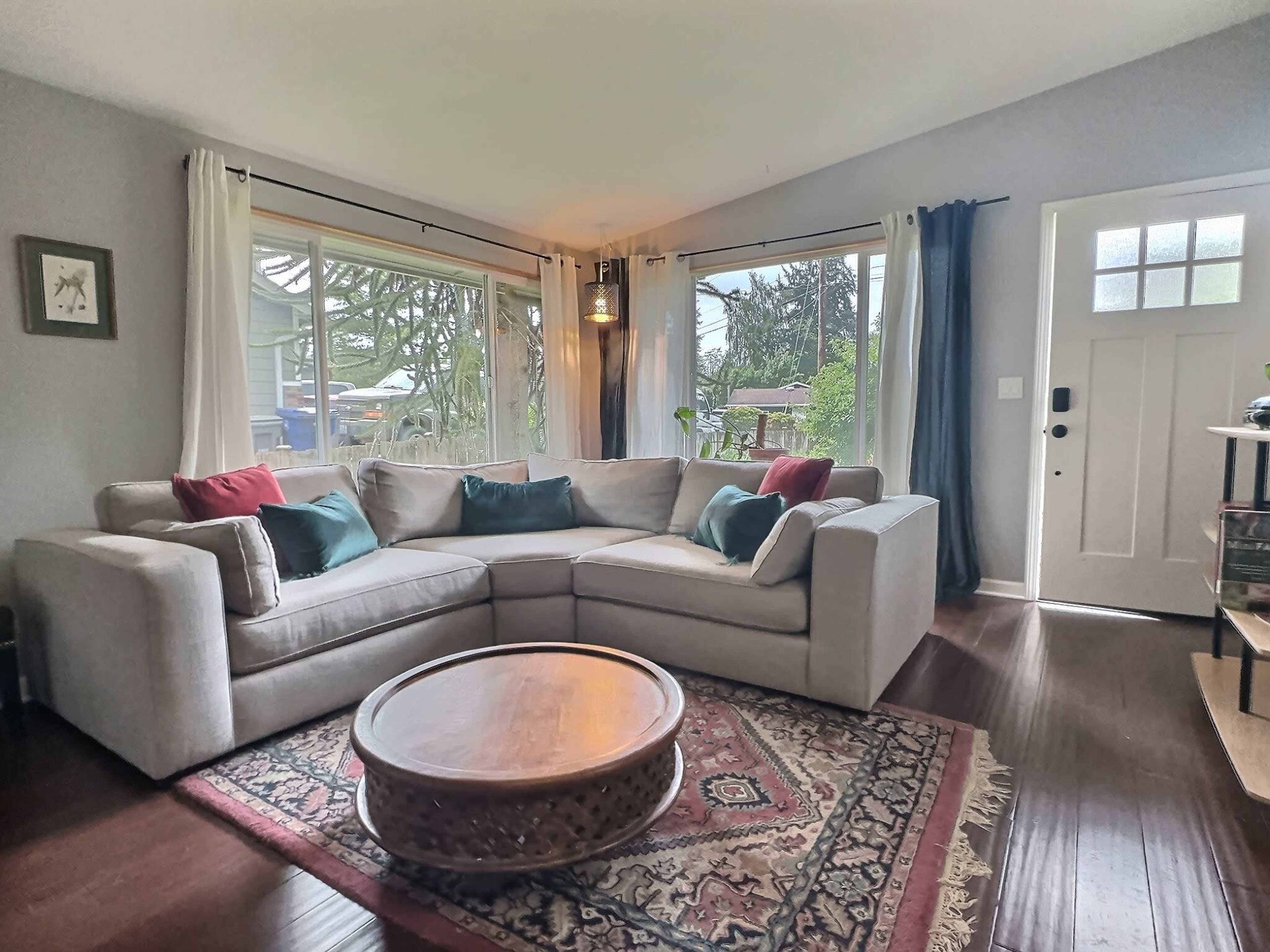 A spacious living room features a large sectional sofa with colorful pillows, a round coffee table, and windows letting in natural light.