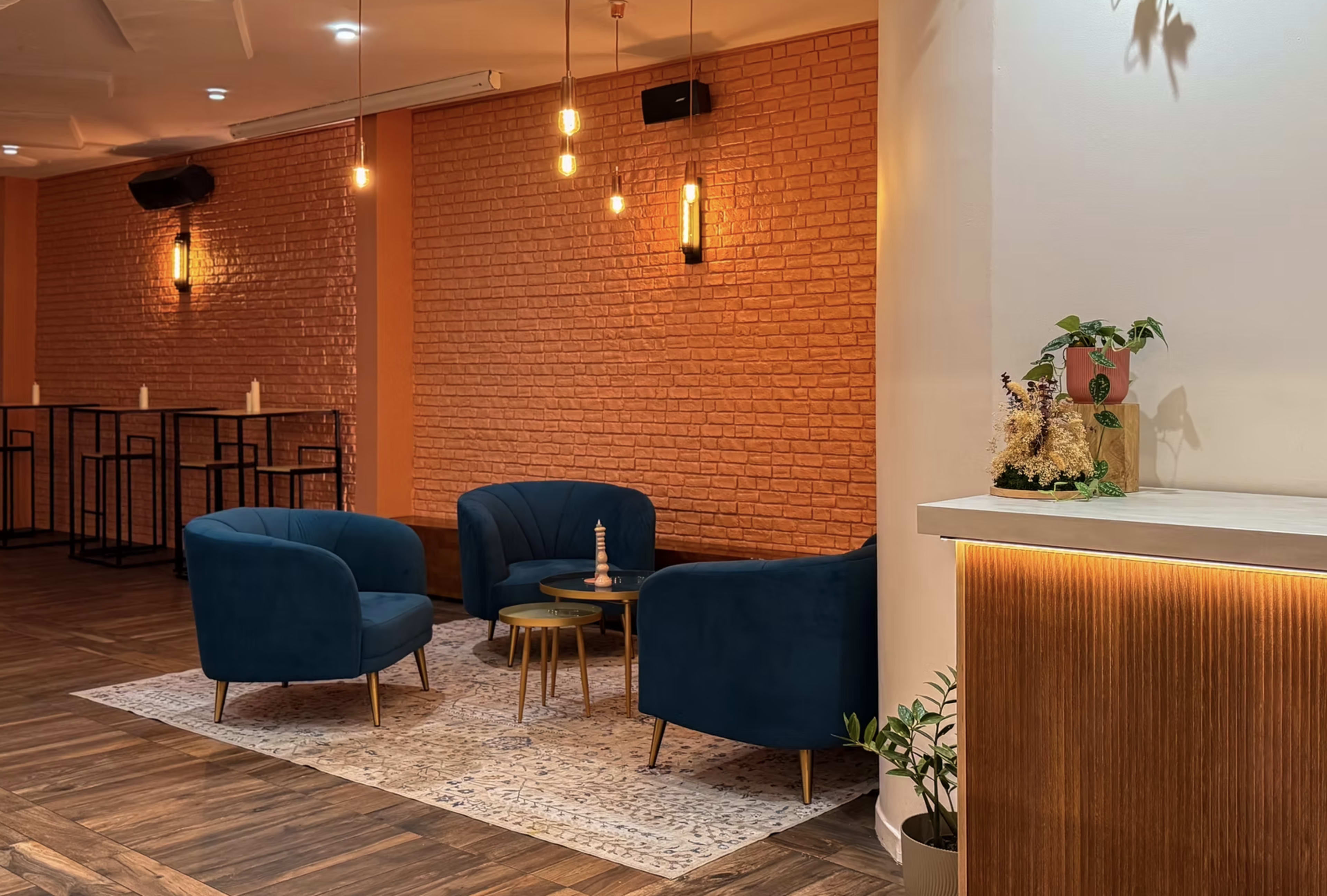 The image shows a modern lounge area featuring three blue chairs arranged around a small coffee table on a patterned rug, with a brick wall and warm ambient lighting in the background.