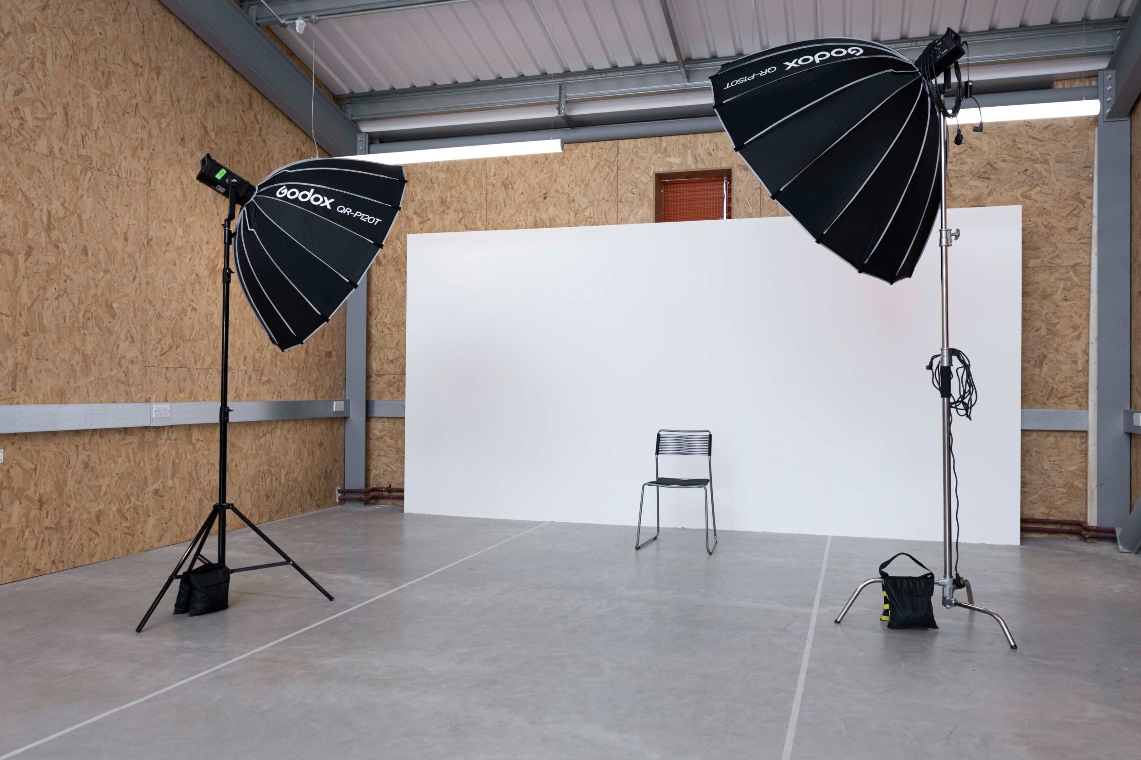 The image shows a photography studio with two large softbox lights facing an empty chair in front of a plain white backdrop.