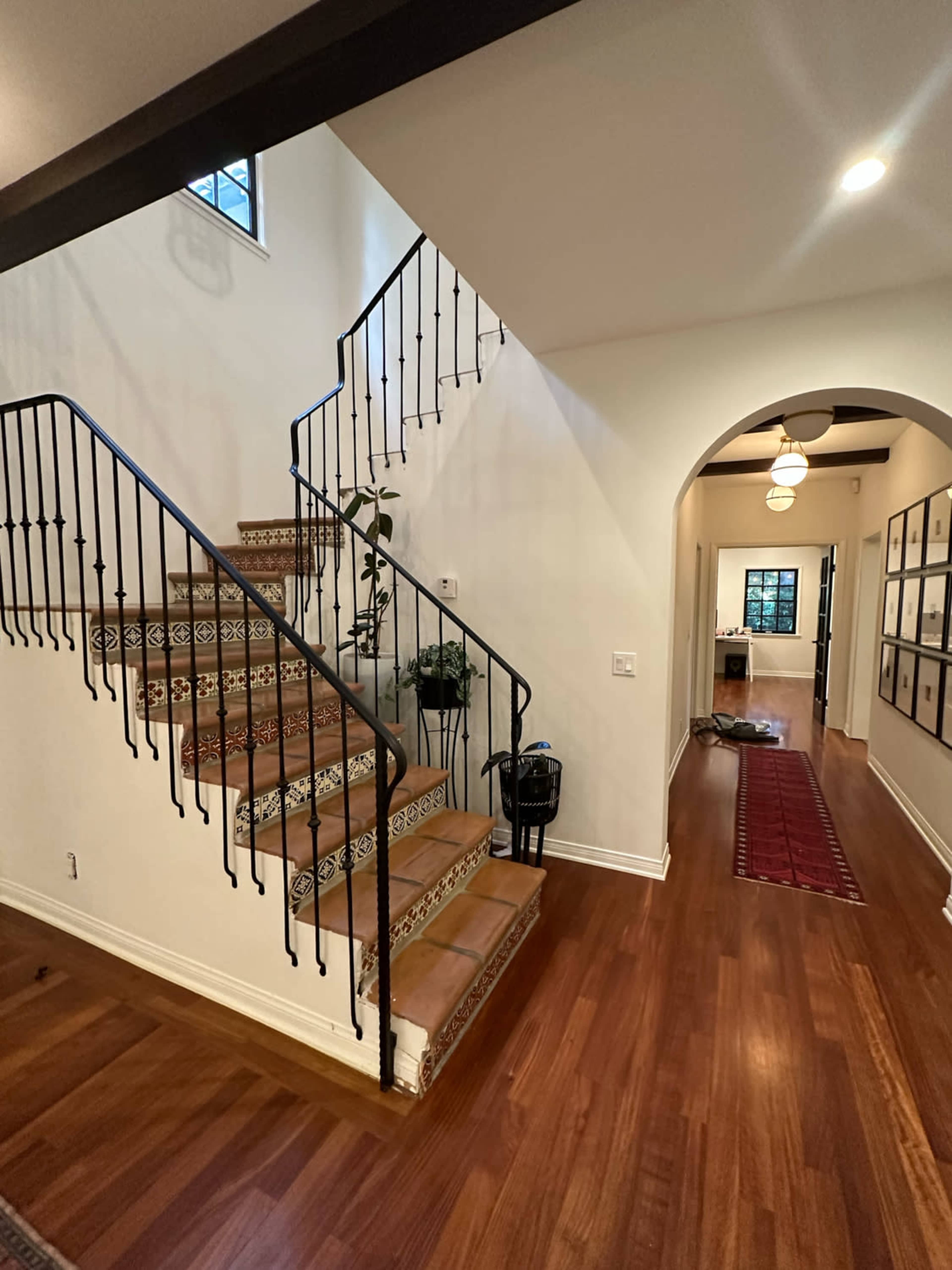 The image shows a brightly lit interior featuring a staircase with decorative tiles, leading to an upper level, alongside a hallway with framed pictures and a runner rug.