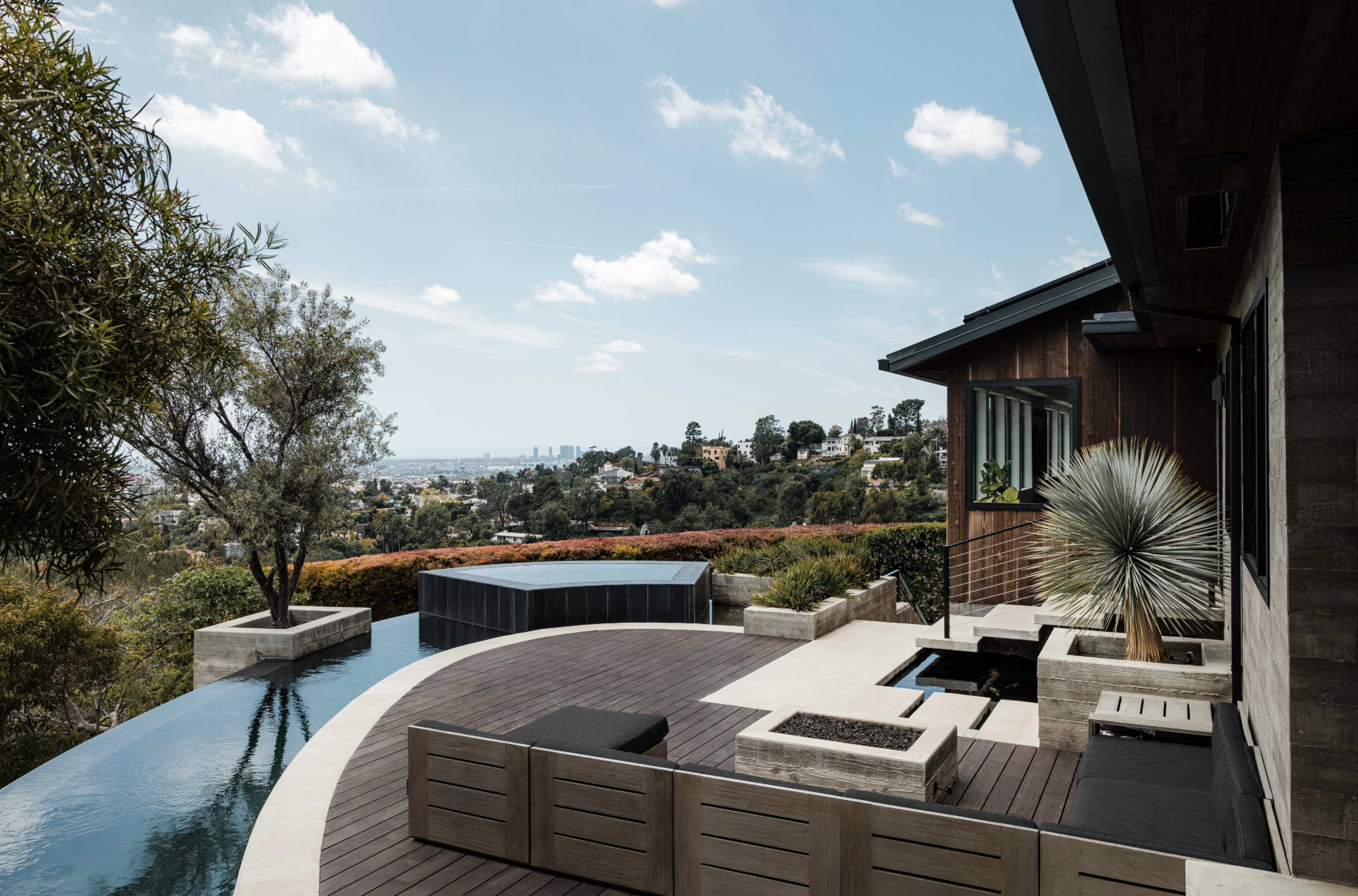 The image shows a modern architectural outdoor space with a pool, wooden decking, and lush landscaping overlooking a distant city skyline.