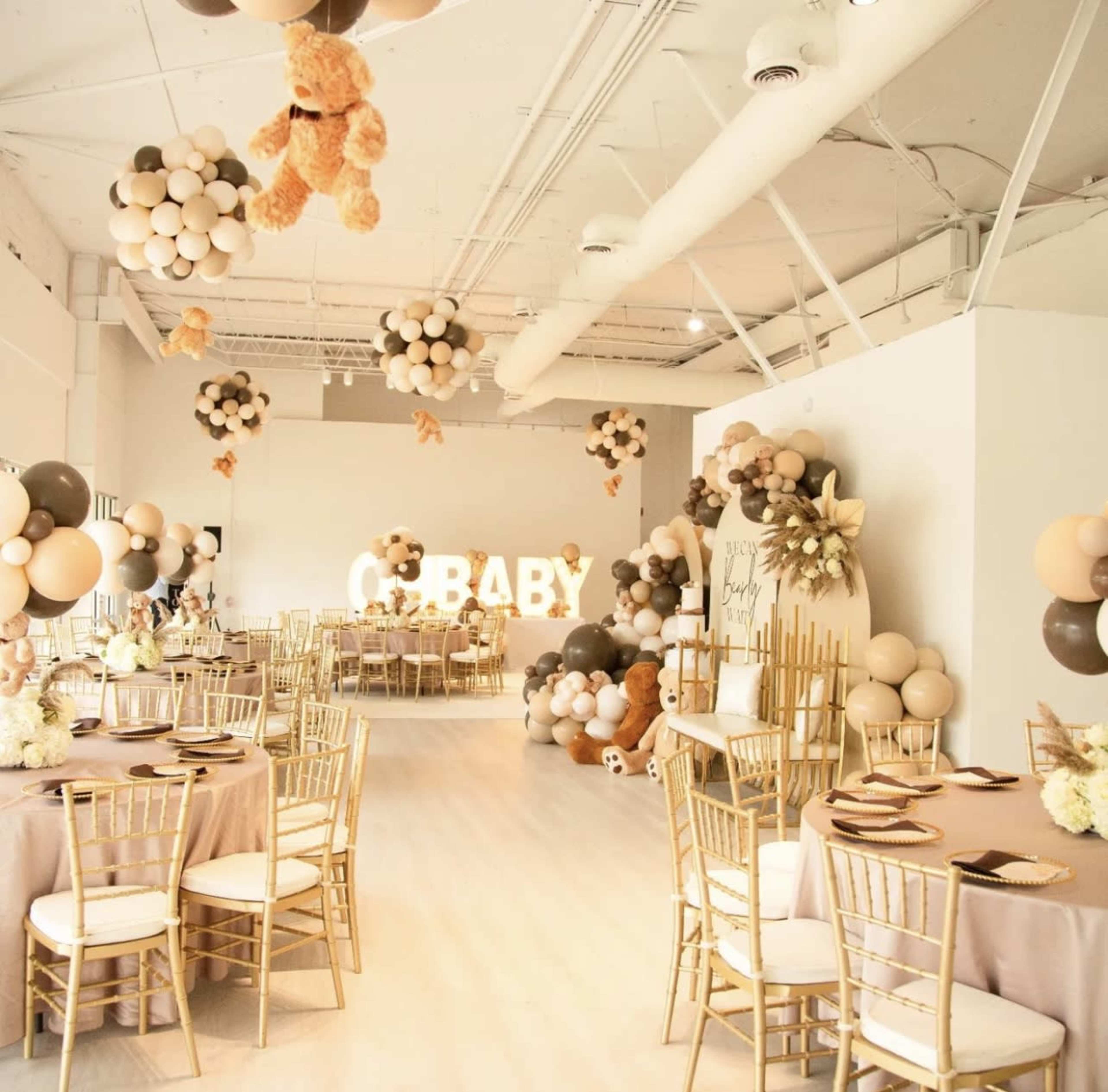 A celebratory room decorated for a baby shower, featuring tables with gold chairs, balloons in various colors, and a "BABY" light display.