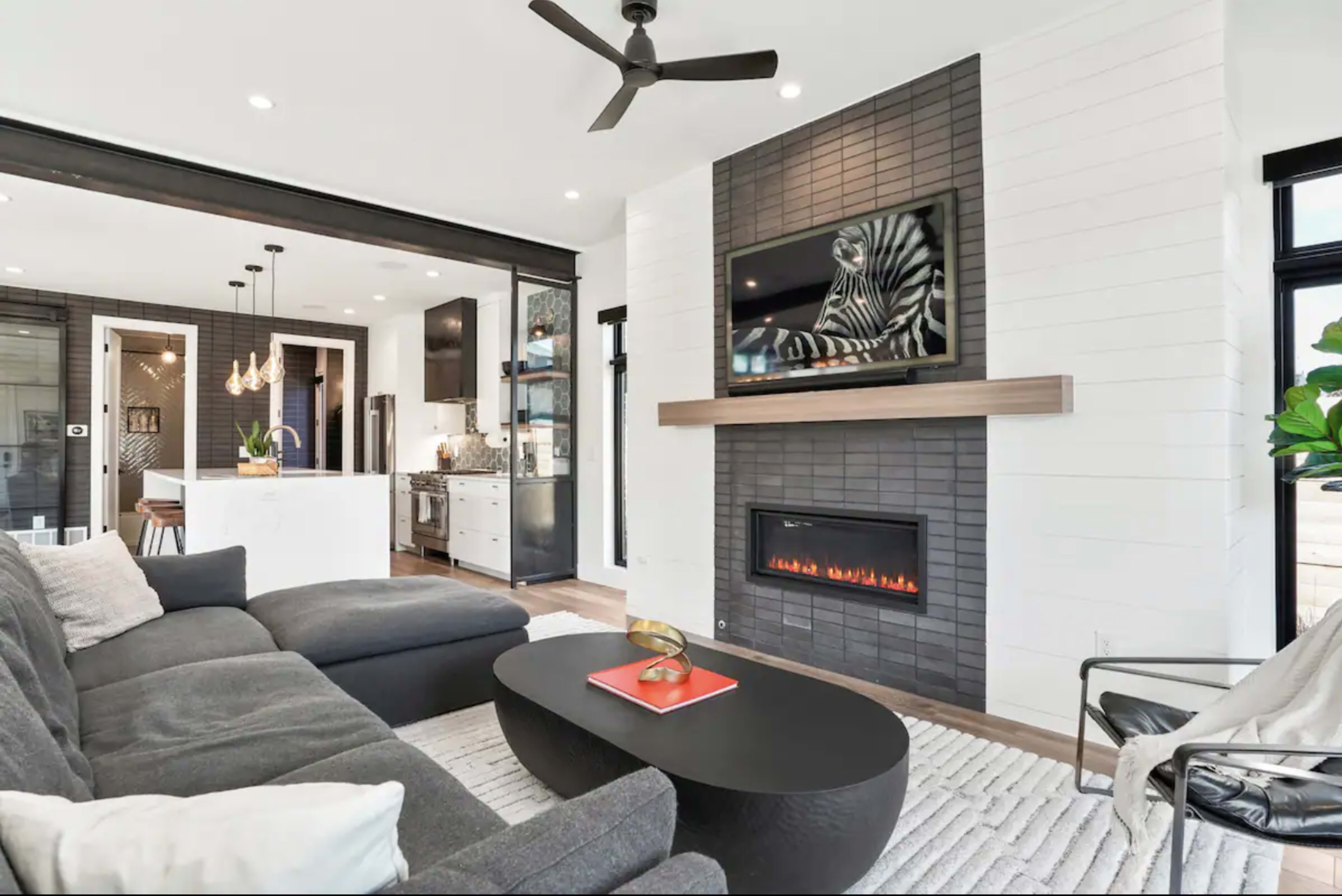 A modern living room with a sectional sofa, an elegant coffee table, a wall-mounted television above a sleek fireplace, and an open view of an adjacent kitchen.