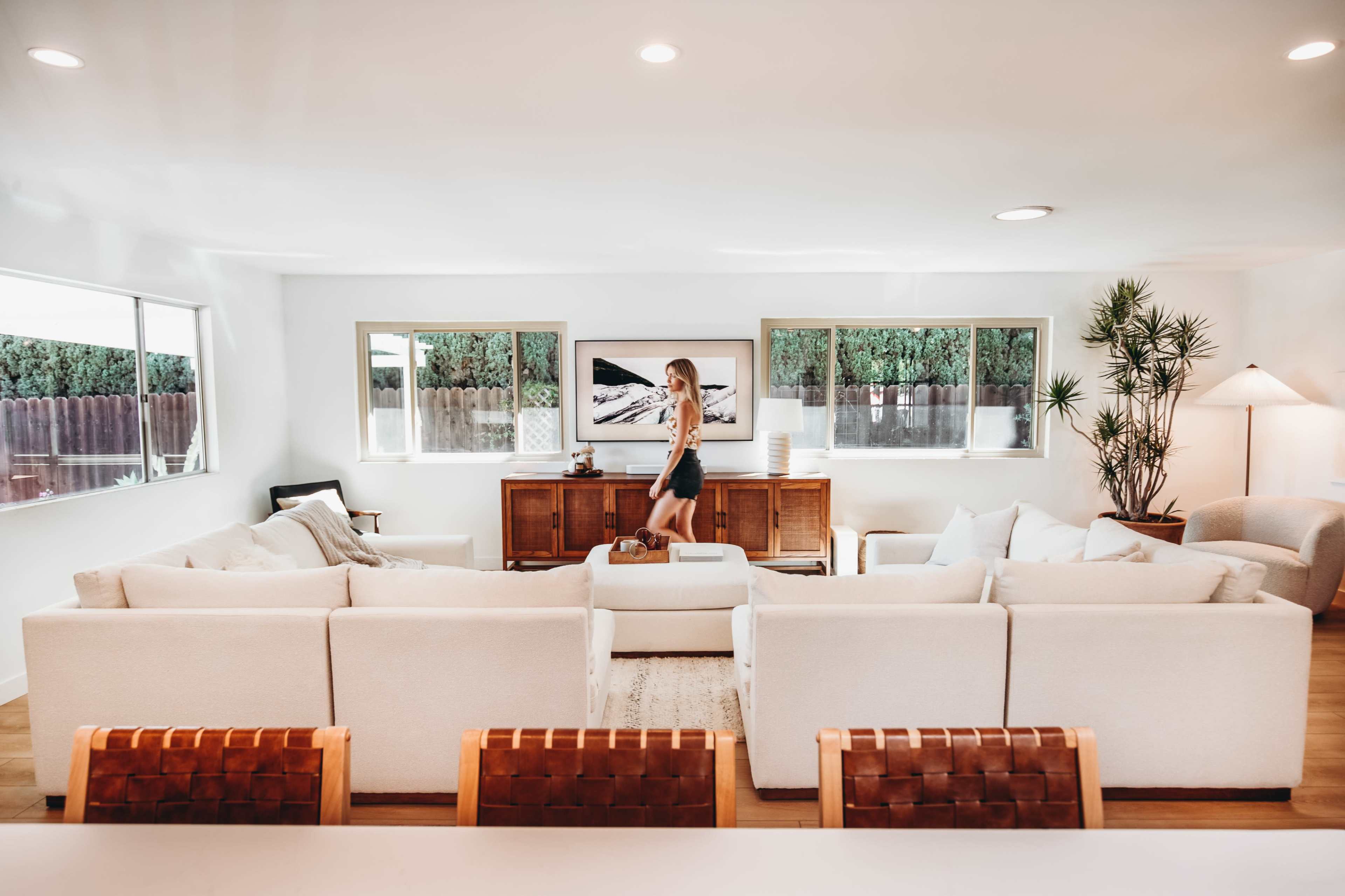 A woman stands near a wooden sideboard in a bright, spacious living room furnished with white couches and large windows.
