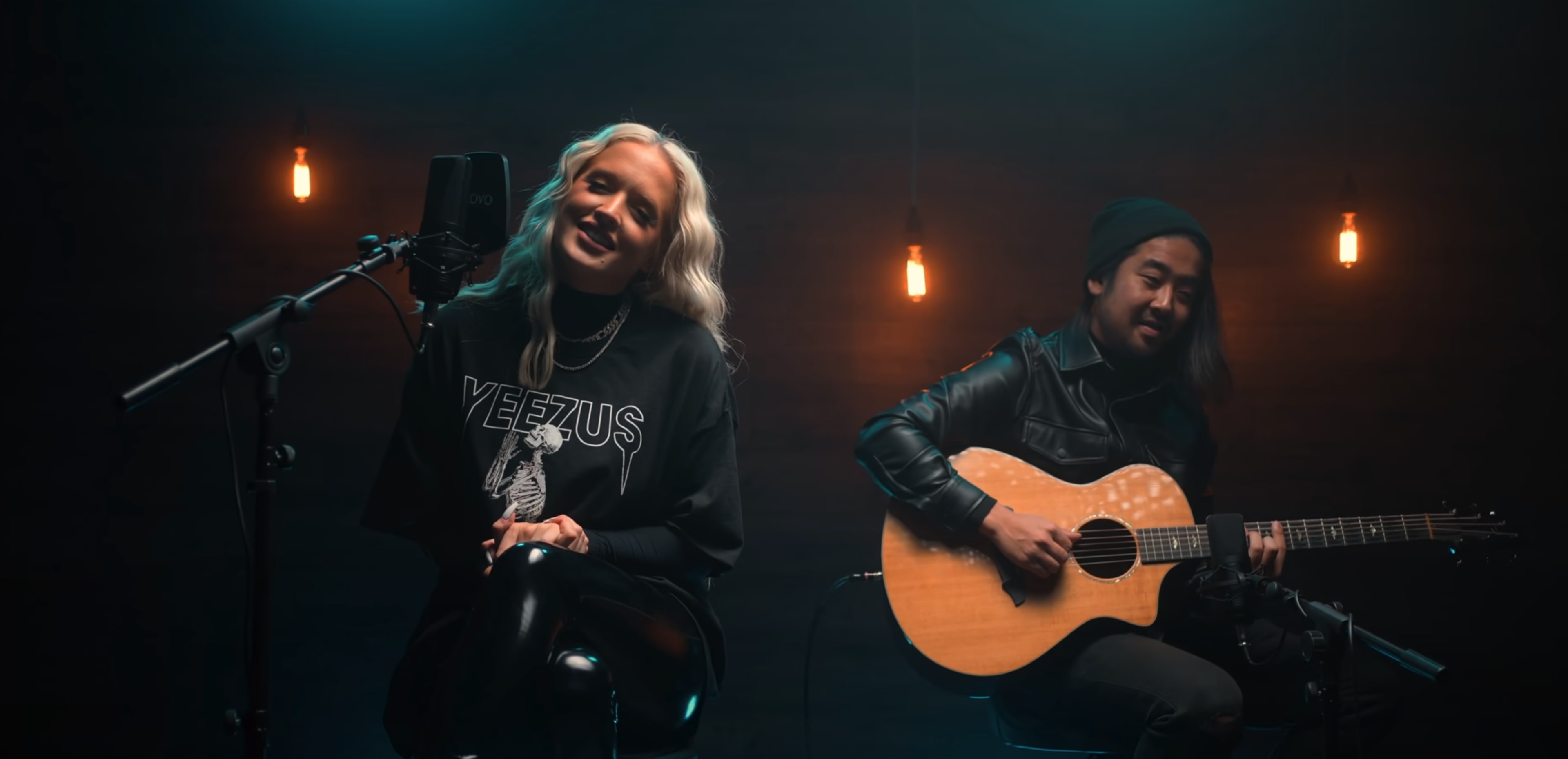 A woman sings into a microphone while a man plays an acoustic guitar beside her in a dimly lit setting with hanging lights.