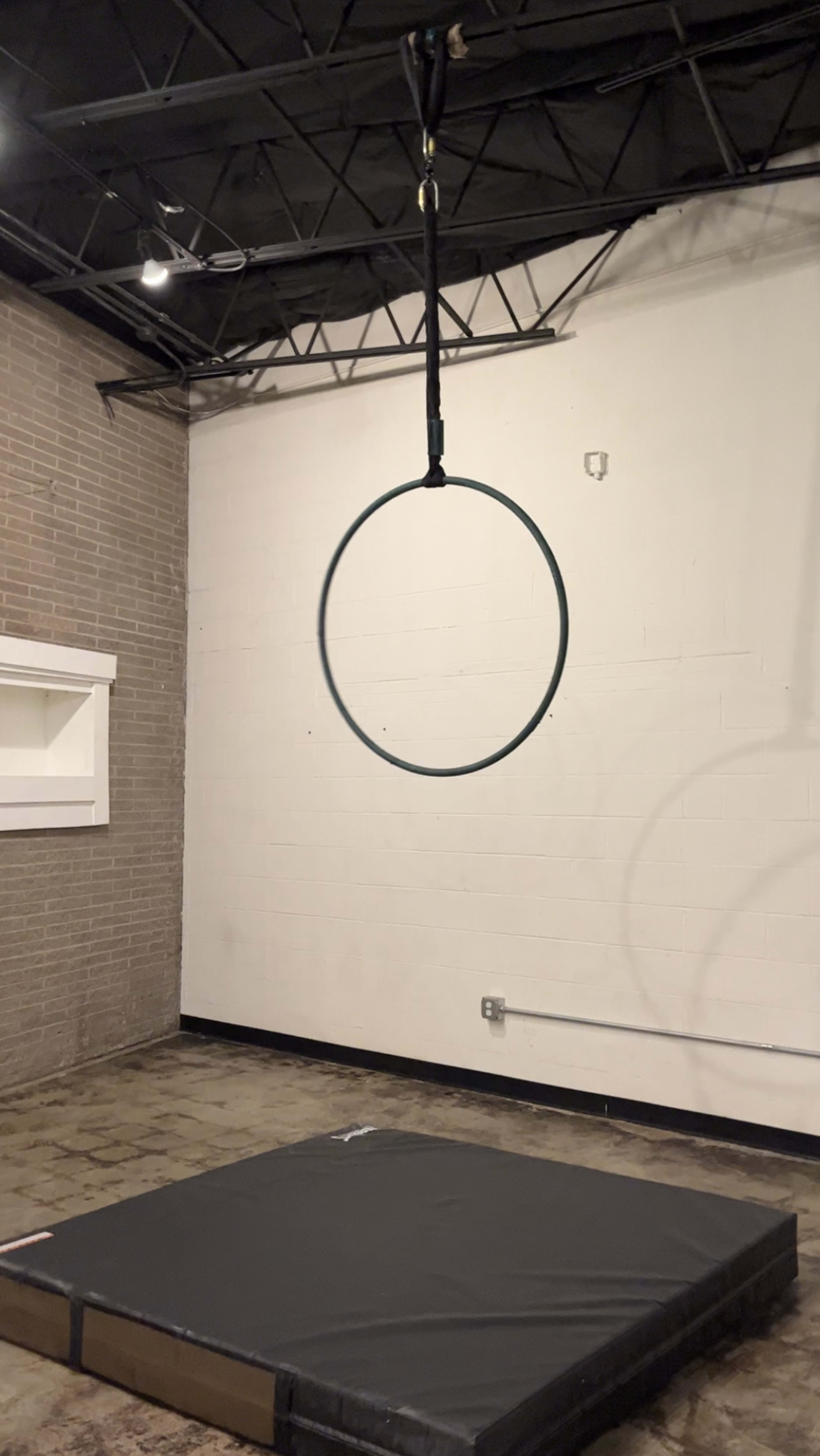 A black aerial hoop hangs from the ceiling above a padded mat in a sparse room.