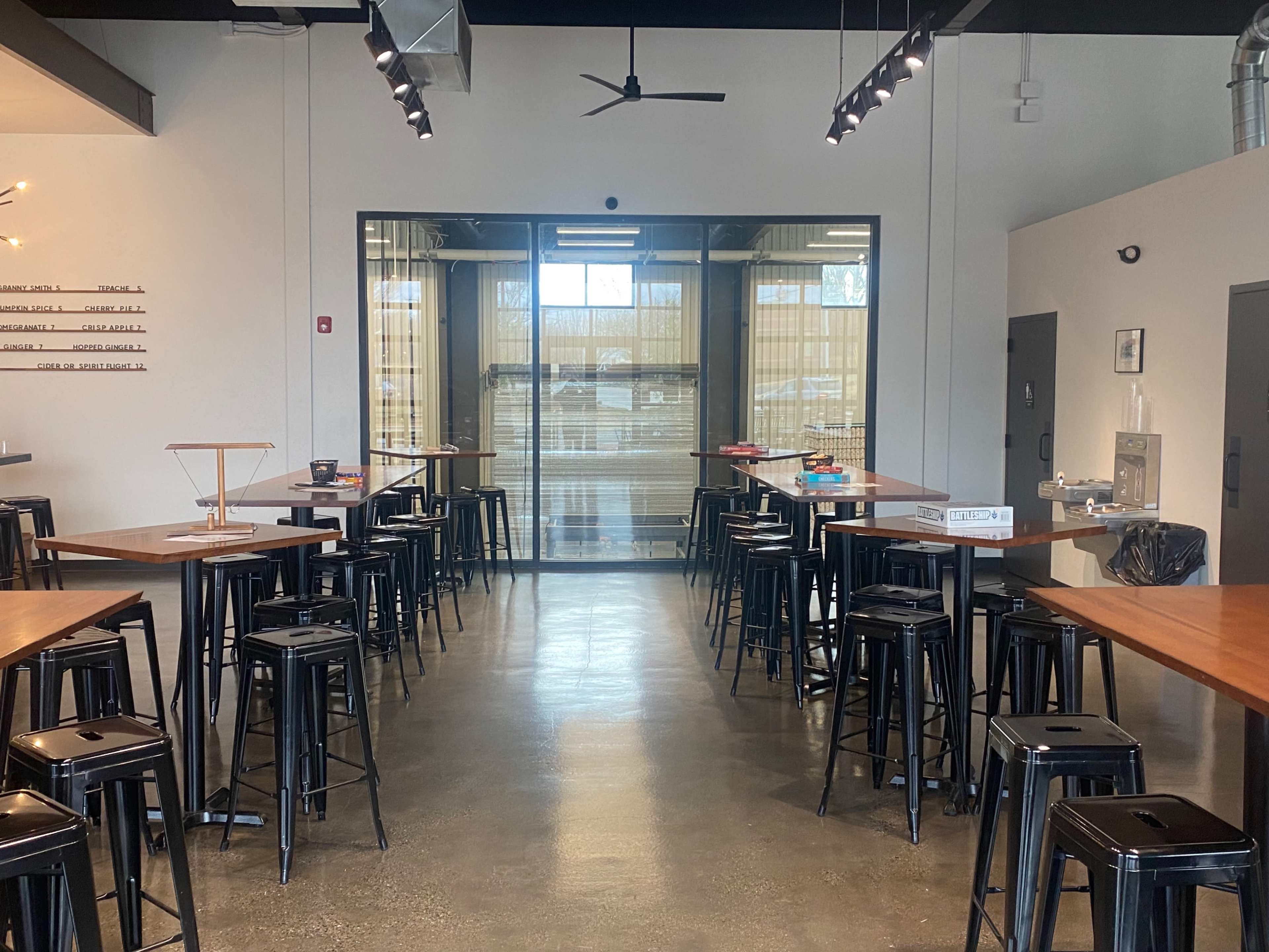 The image shows a modern indoor space with high tables and black stools arranged in rows, leading to a large glass door that provides access to the outside.
