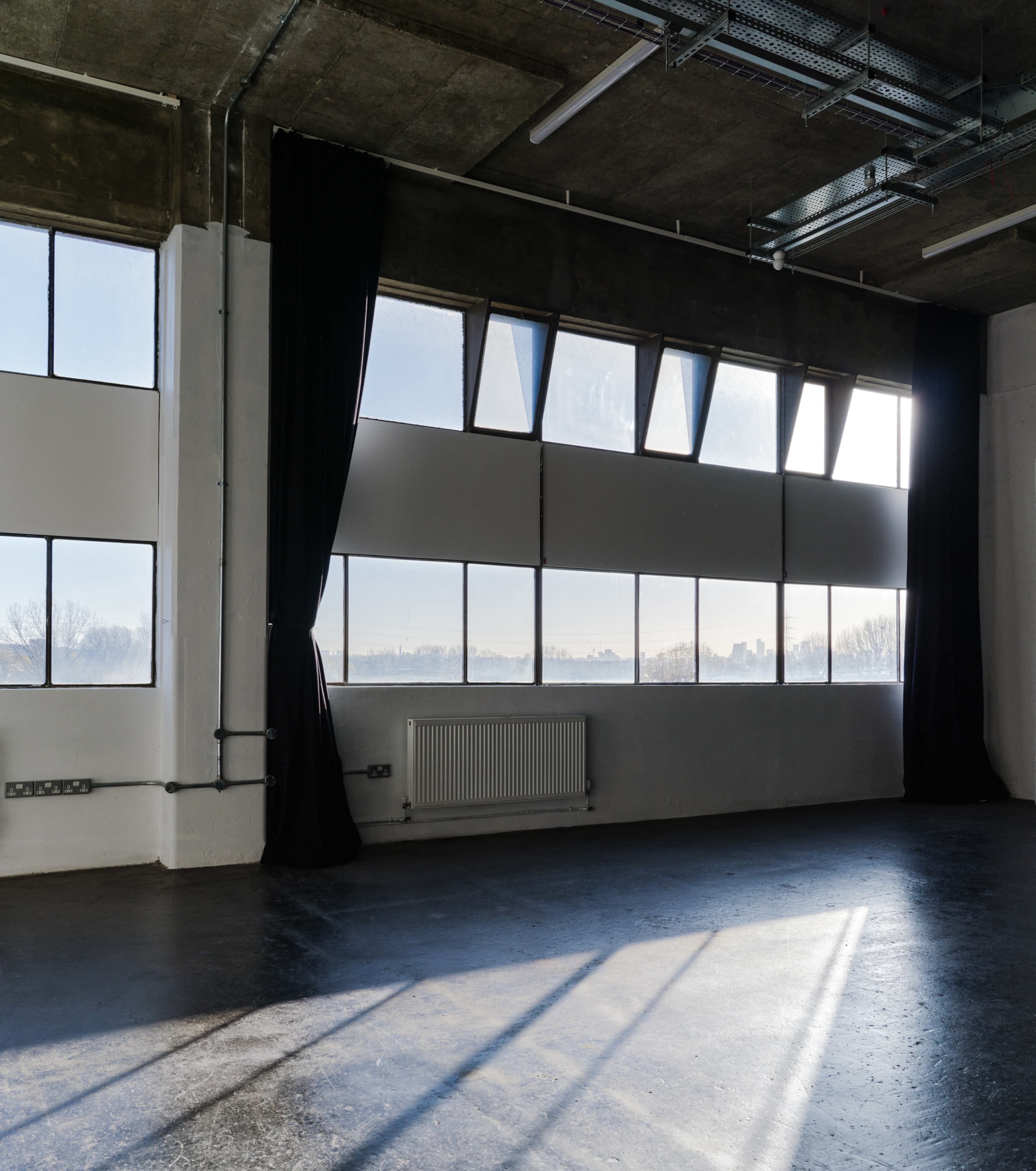 A bright, empty room features large windows with sheer curtains, concrete walls, and a polished floor.