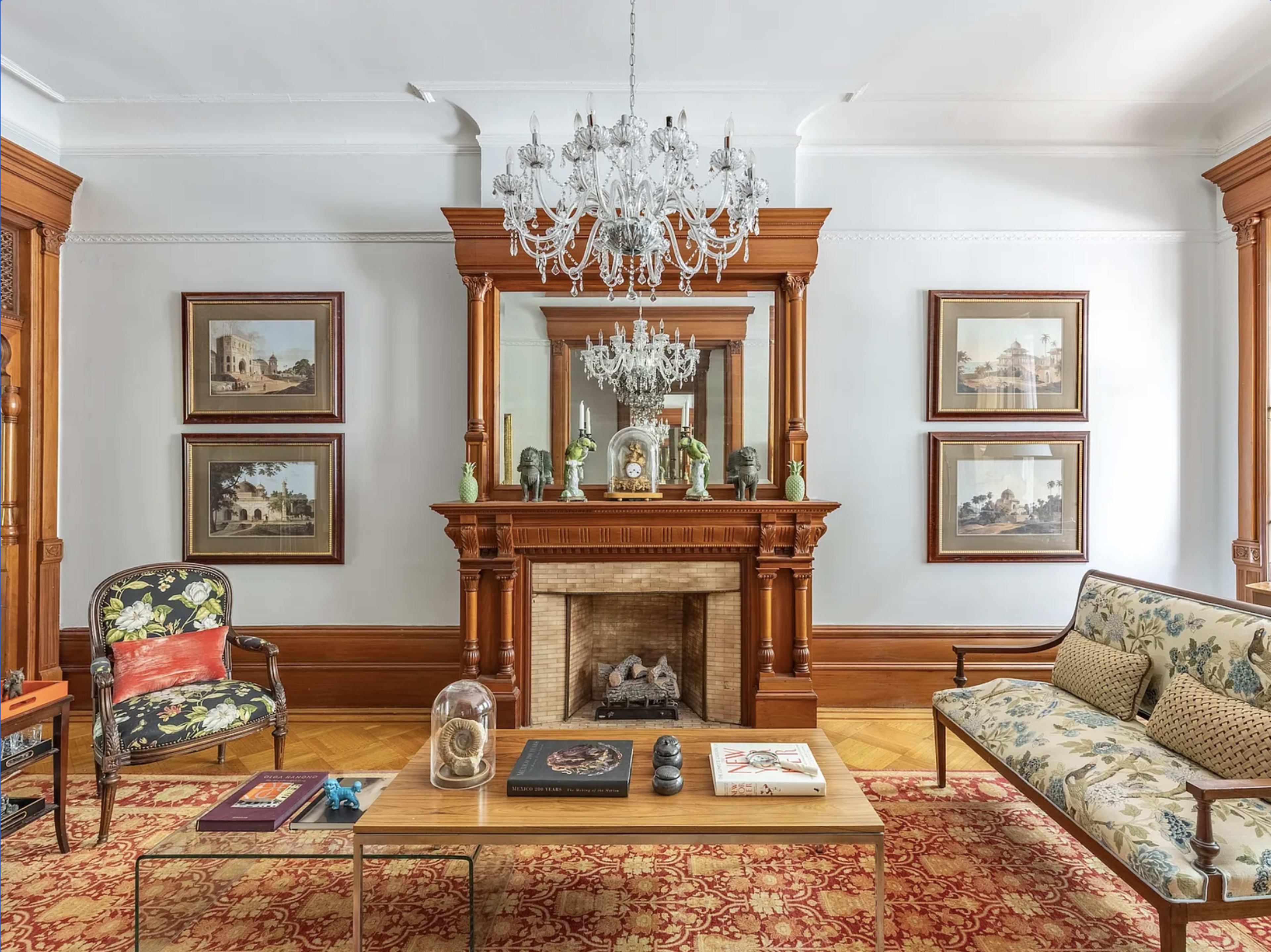 The room features a wooden mantel with a mirror above it, surrounded by three framed artworks, a chandelier overhead, and a mix of upholstered furniture on a patterned rug.