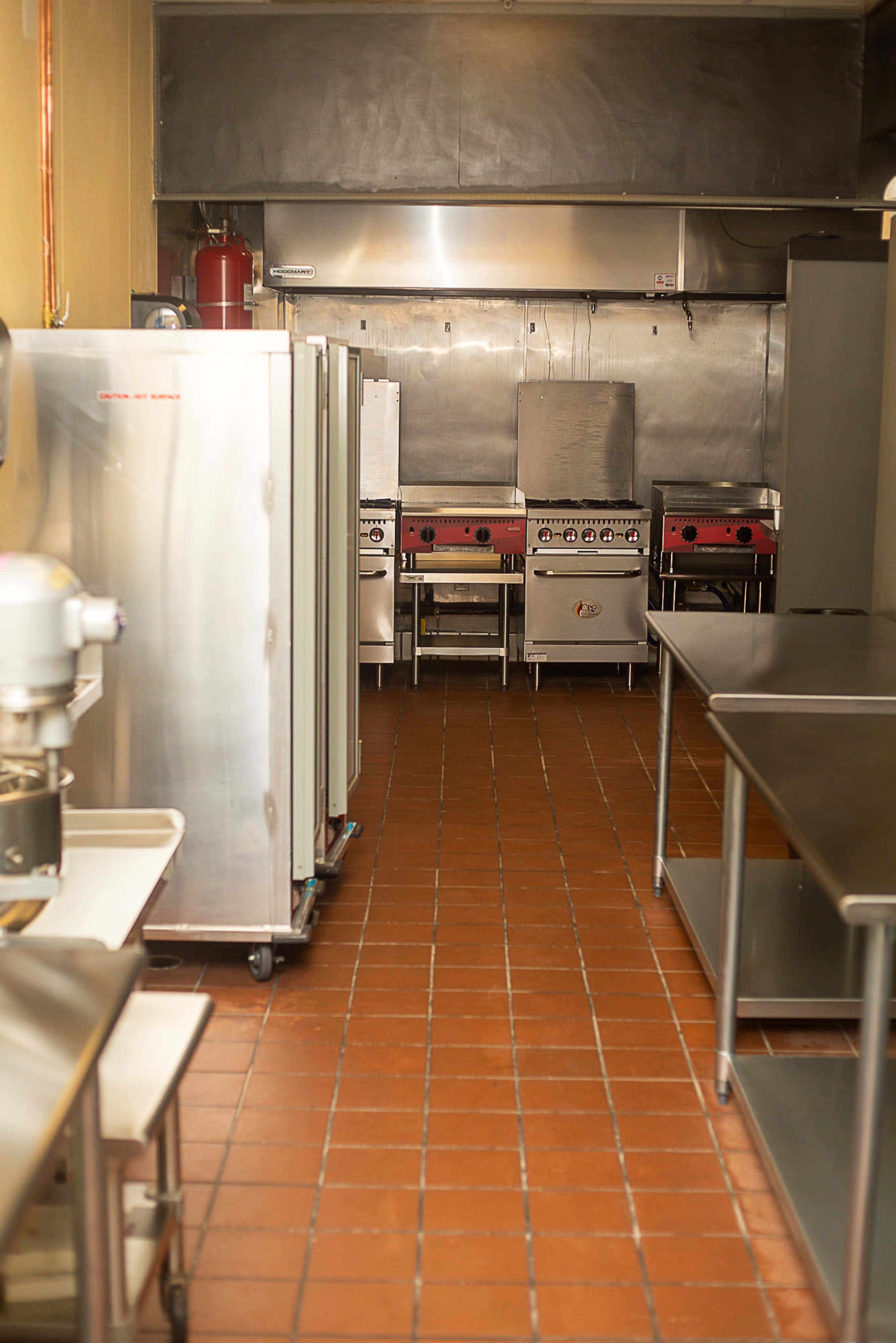 The image shows a commercial kitchen featuring stainless steel appliances, cooking ranges, and work tables set against tiled flooring.