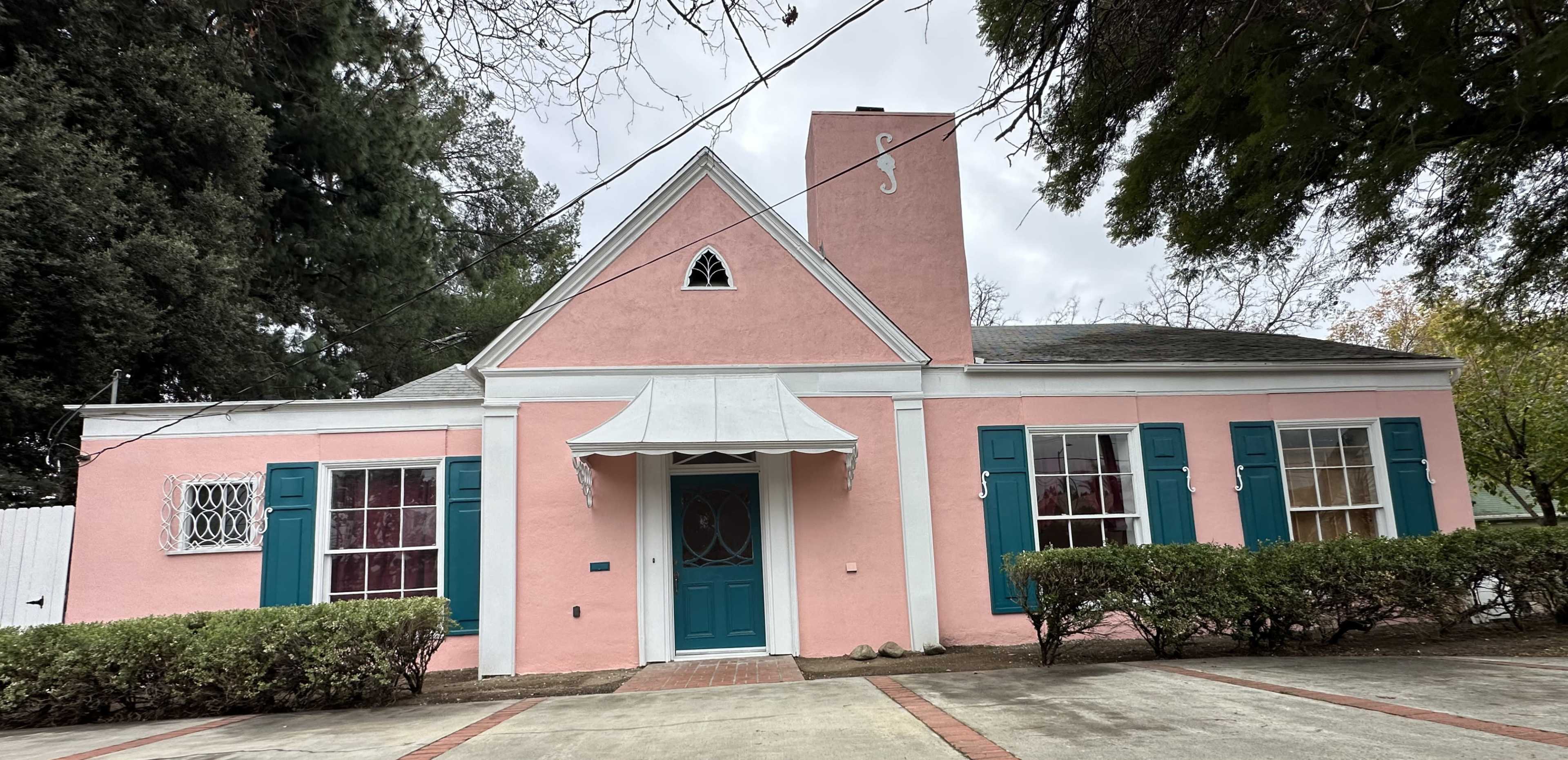 A pink house with teal shutters and a white awning is situated on a landscaped lot.