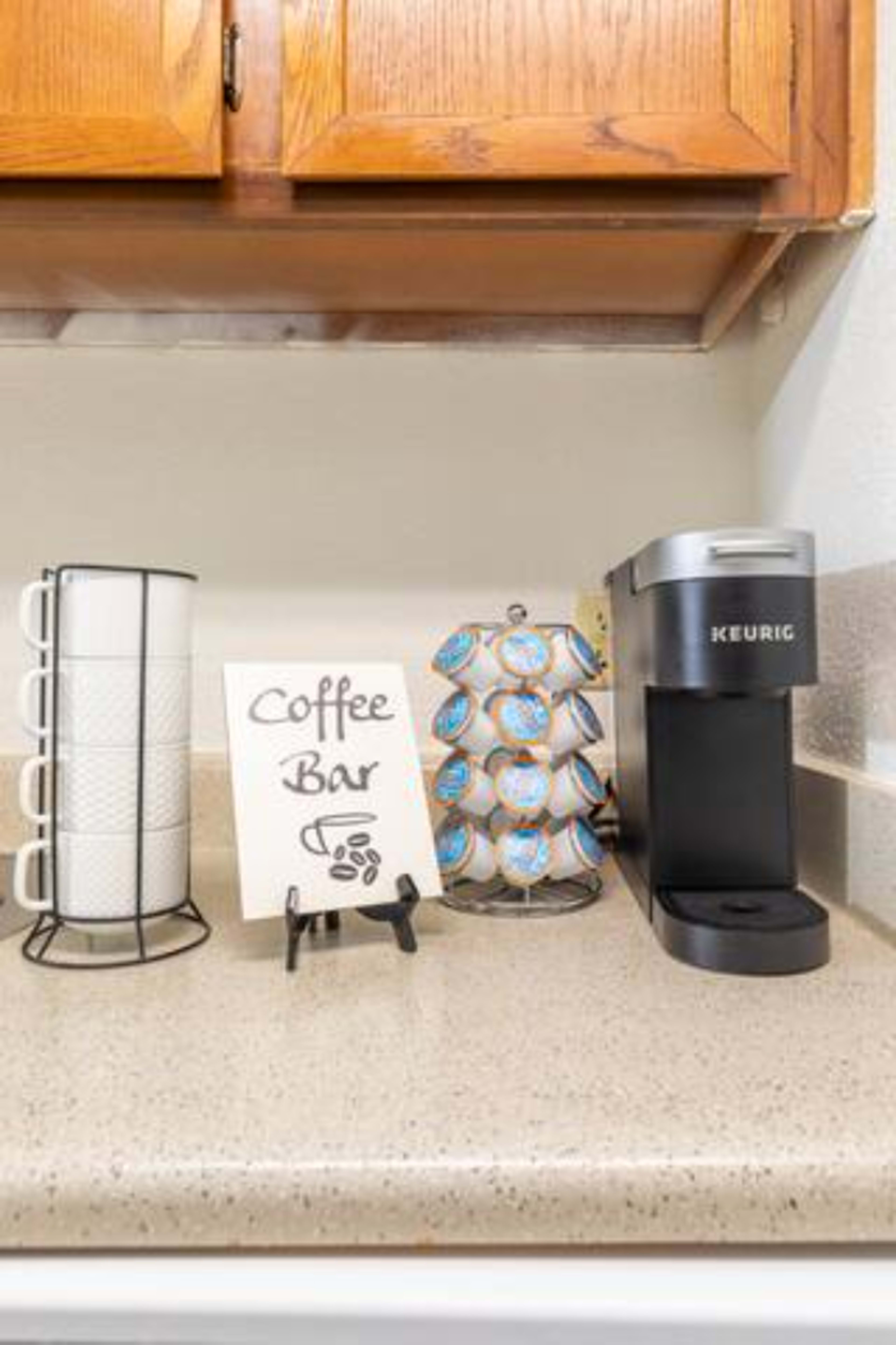 A coffee station featuring a Keurig machine, a rack of coffee pods, and a sign labeled "Coffee Bar" on a countertop beneath wooden cabinets.
