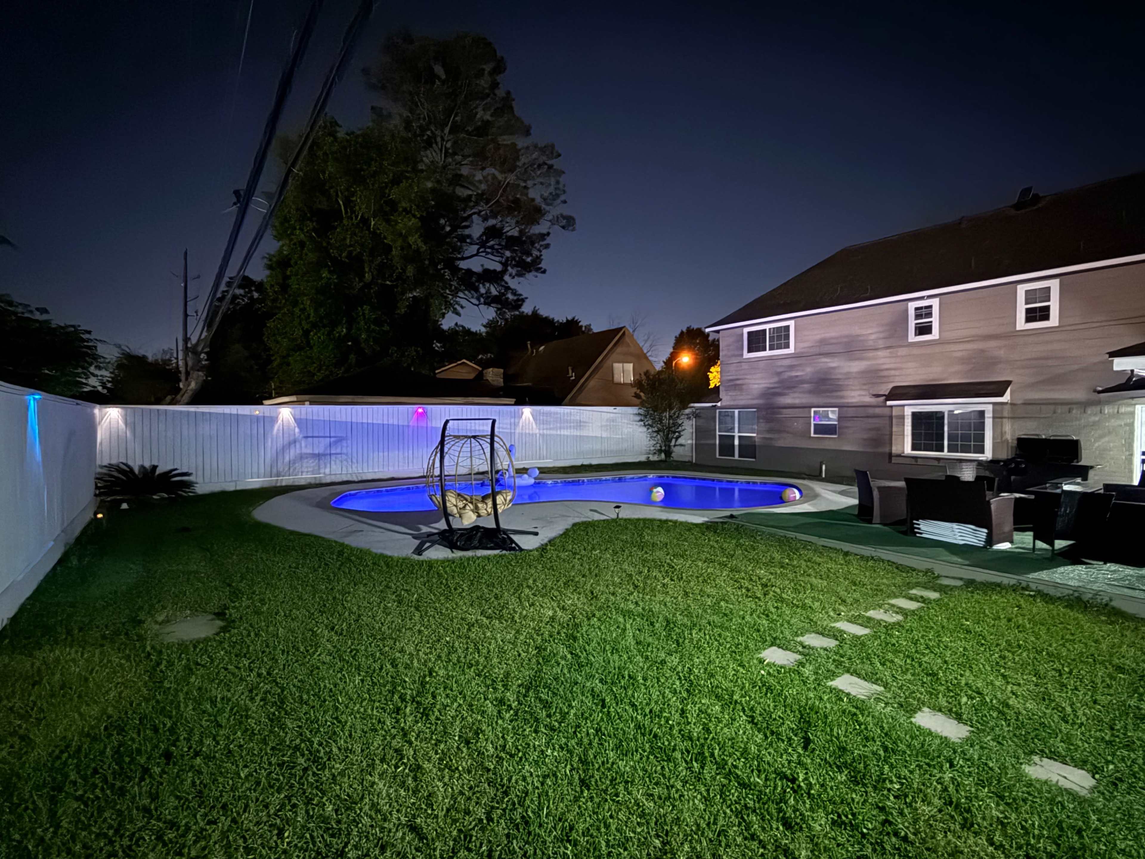 A well-lit backyard features a blue swimming pool, a grassy area, and a seating arrangement beside a house at night.