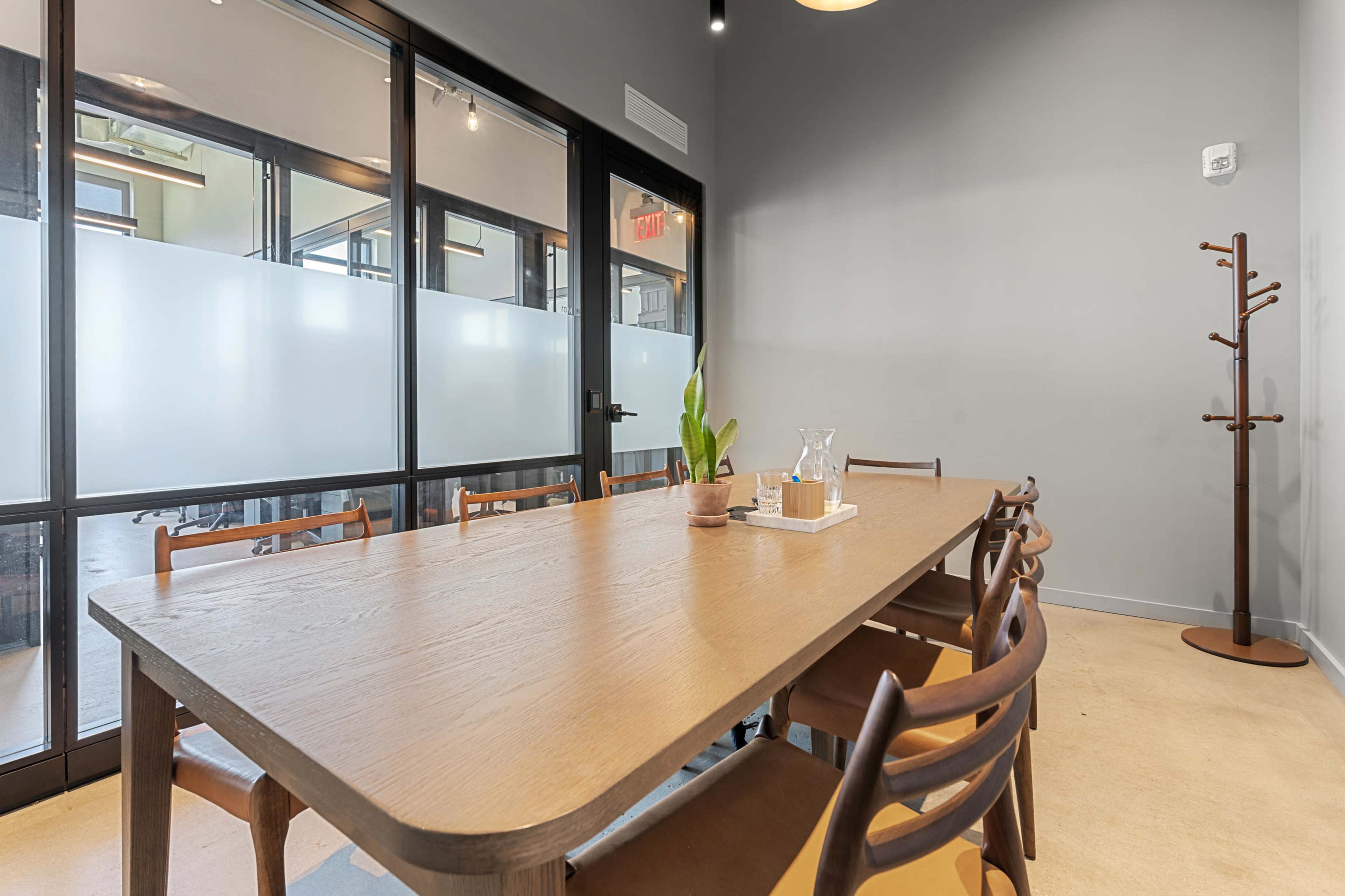 A large wooden table with several chairs is positioned in a modern meeting room, featuring glass walls and a coat rack.