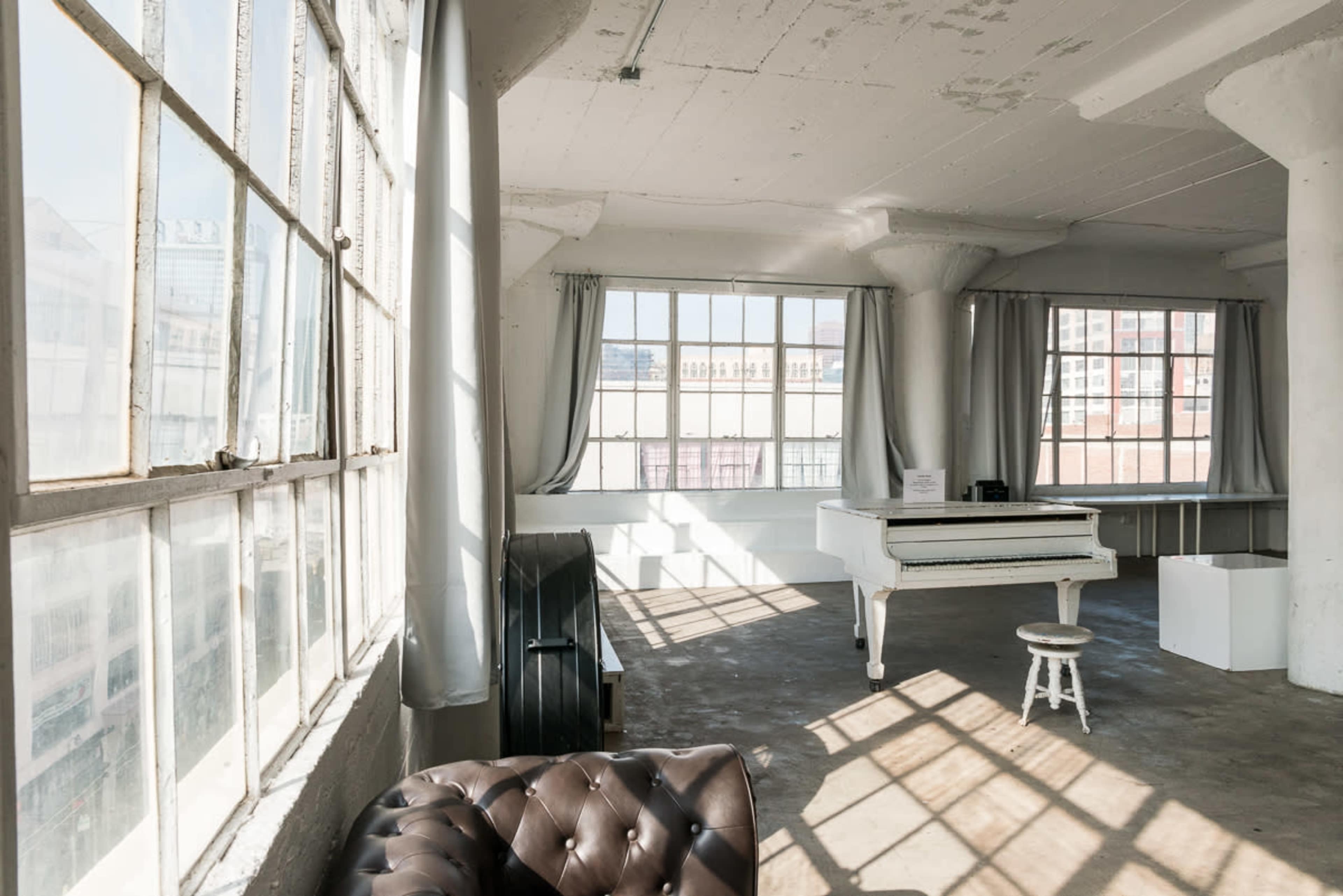 The image shows a spacious interior featuring large windows, a white grand piano, a small stool, and a dark upholstered couch against a light background.