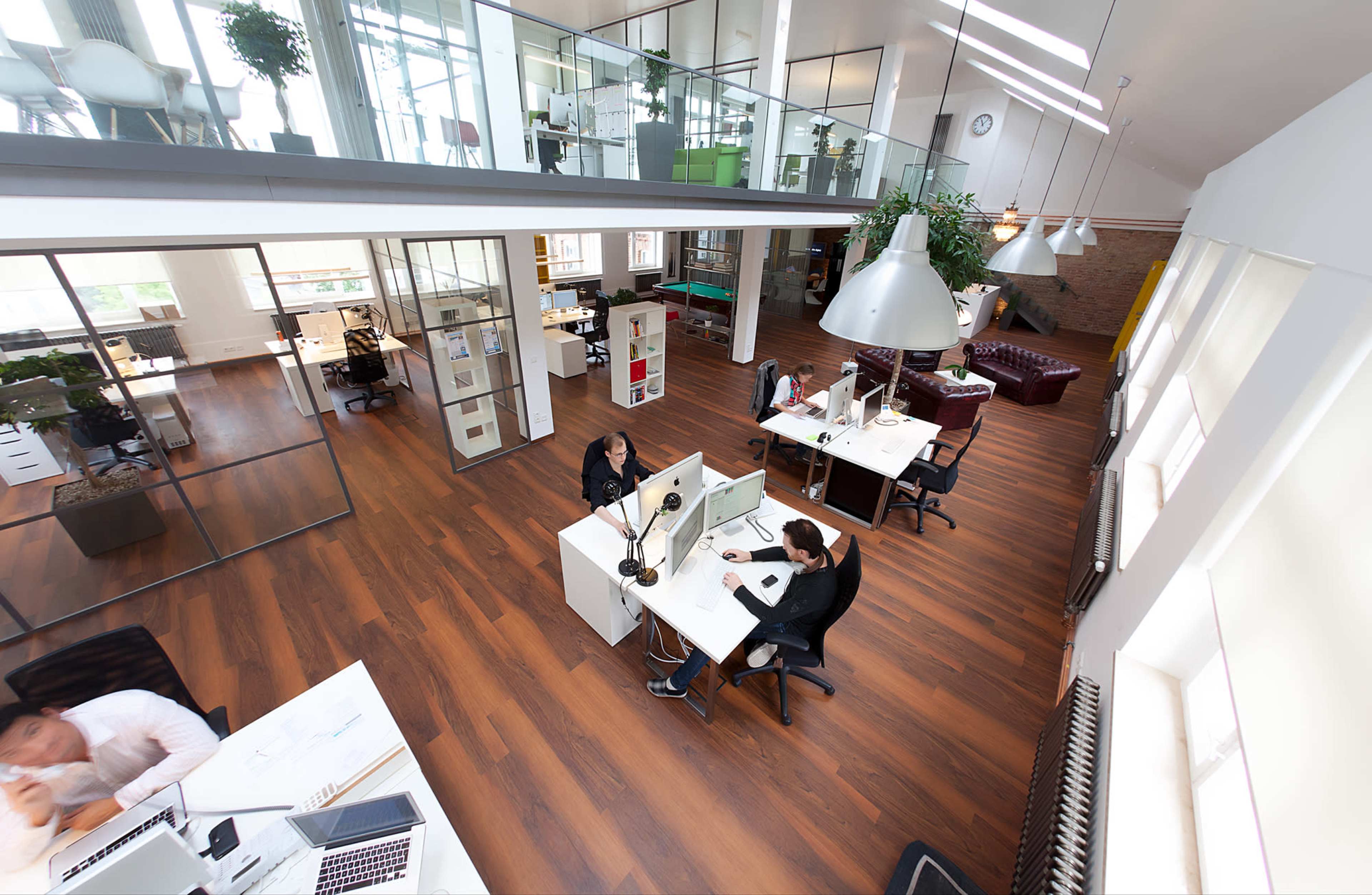 The image shows a modern office space with several workstations, large windows, and a mix of open and enclosed areas.