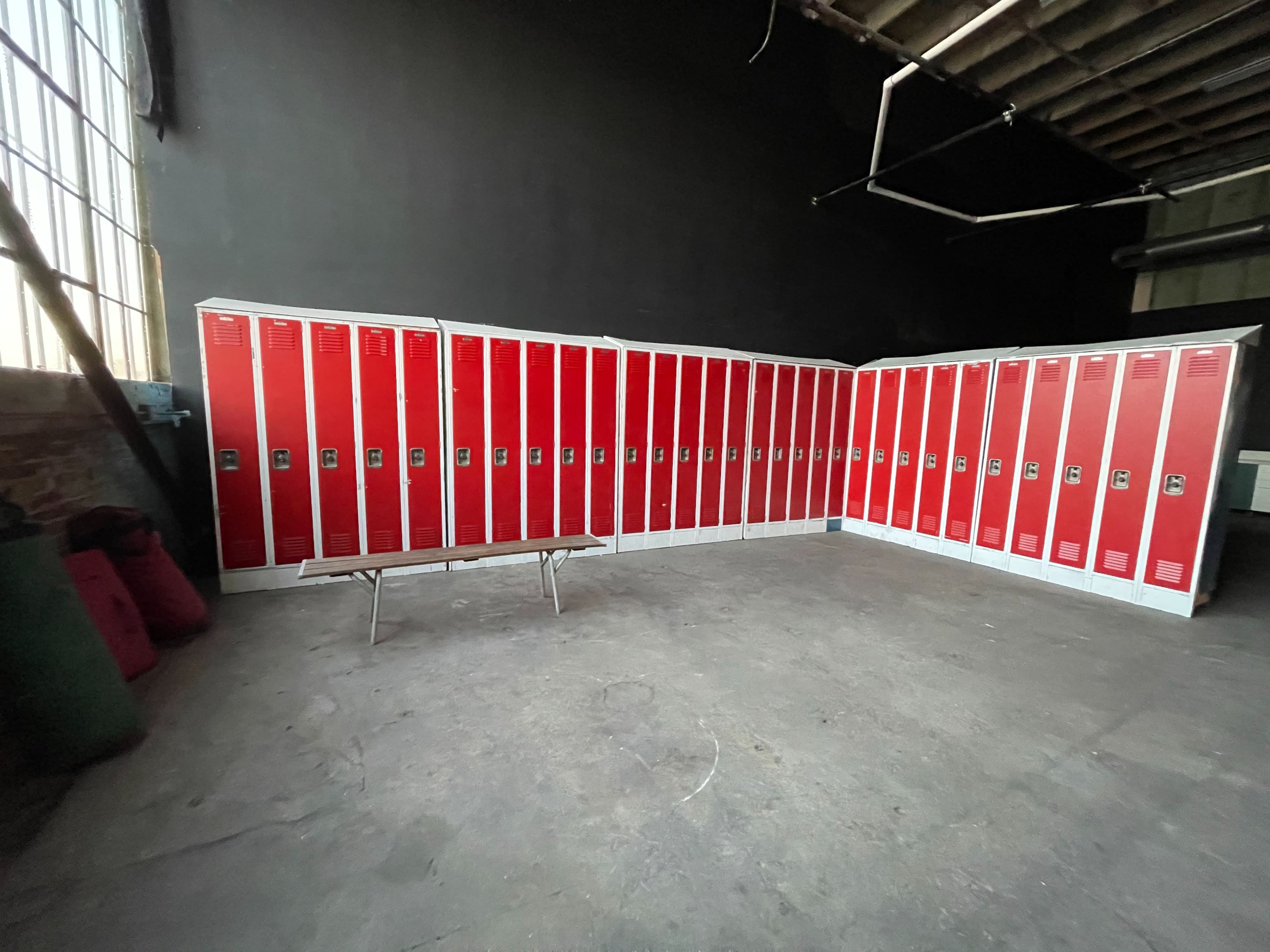 The image shows a row of red lockers arranged in a corner of a spacious, industrial room with a concrete floor.