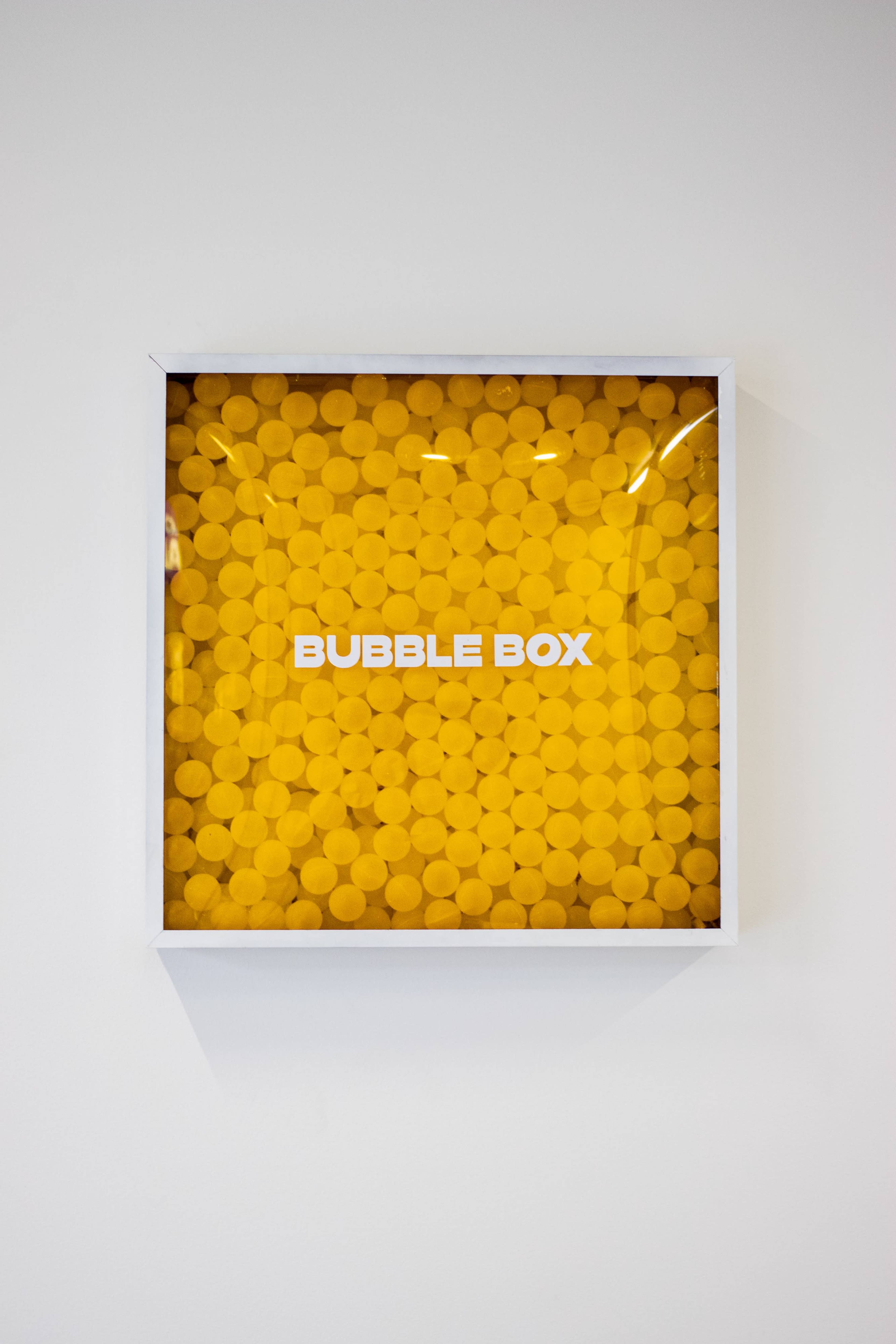 A display box filled with yellow circular foam pieces is mounted on a white wall.