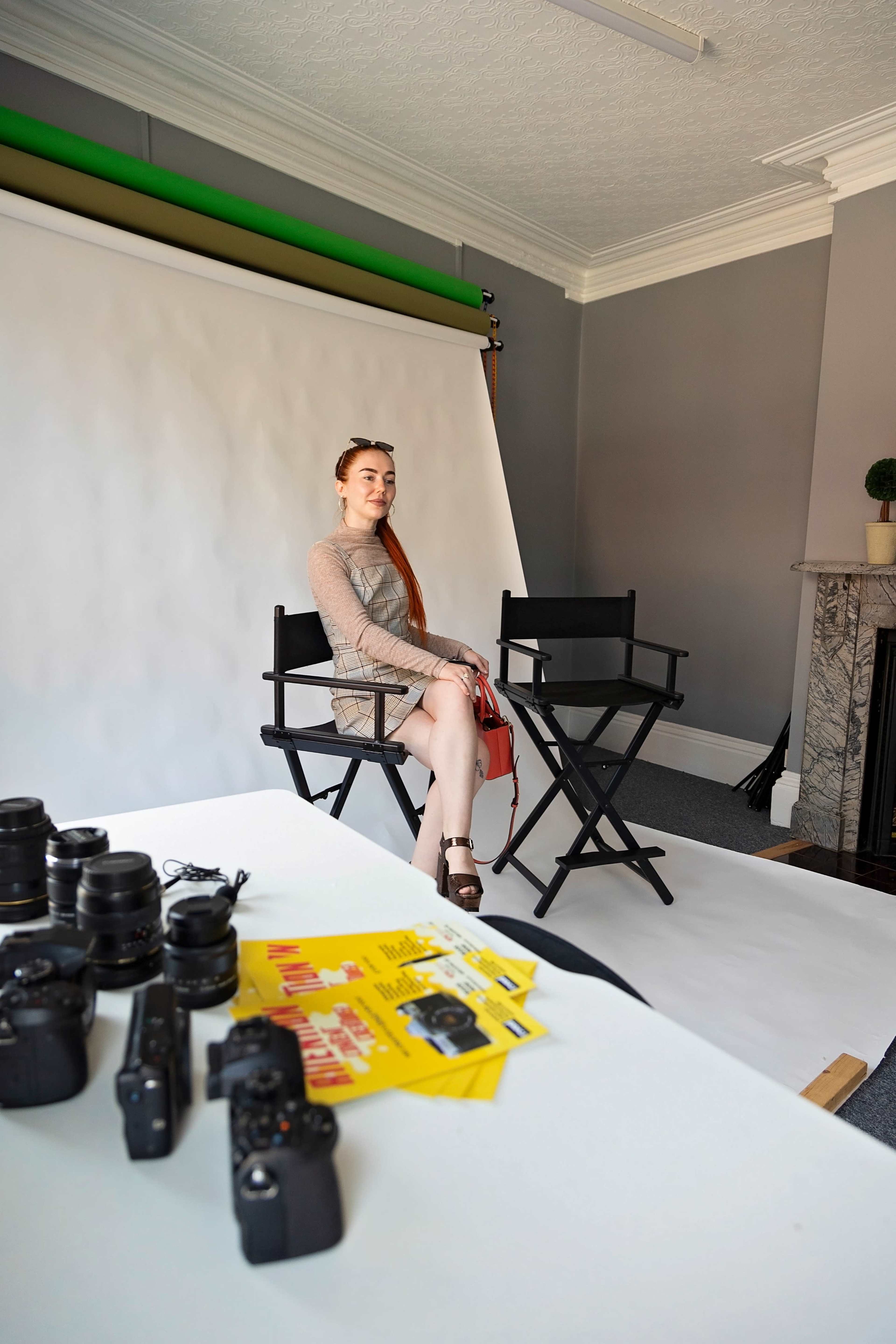 A model sits between two director's chairs in a photography studio, surrounded by cameras and promotional materials on a table.