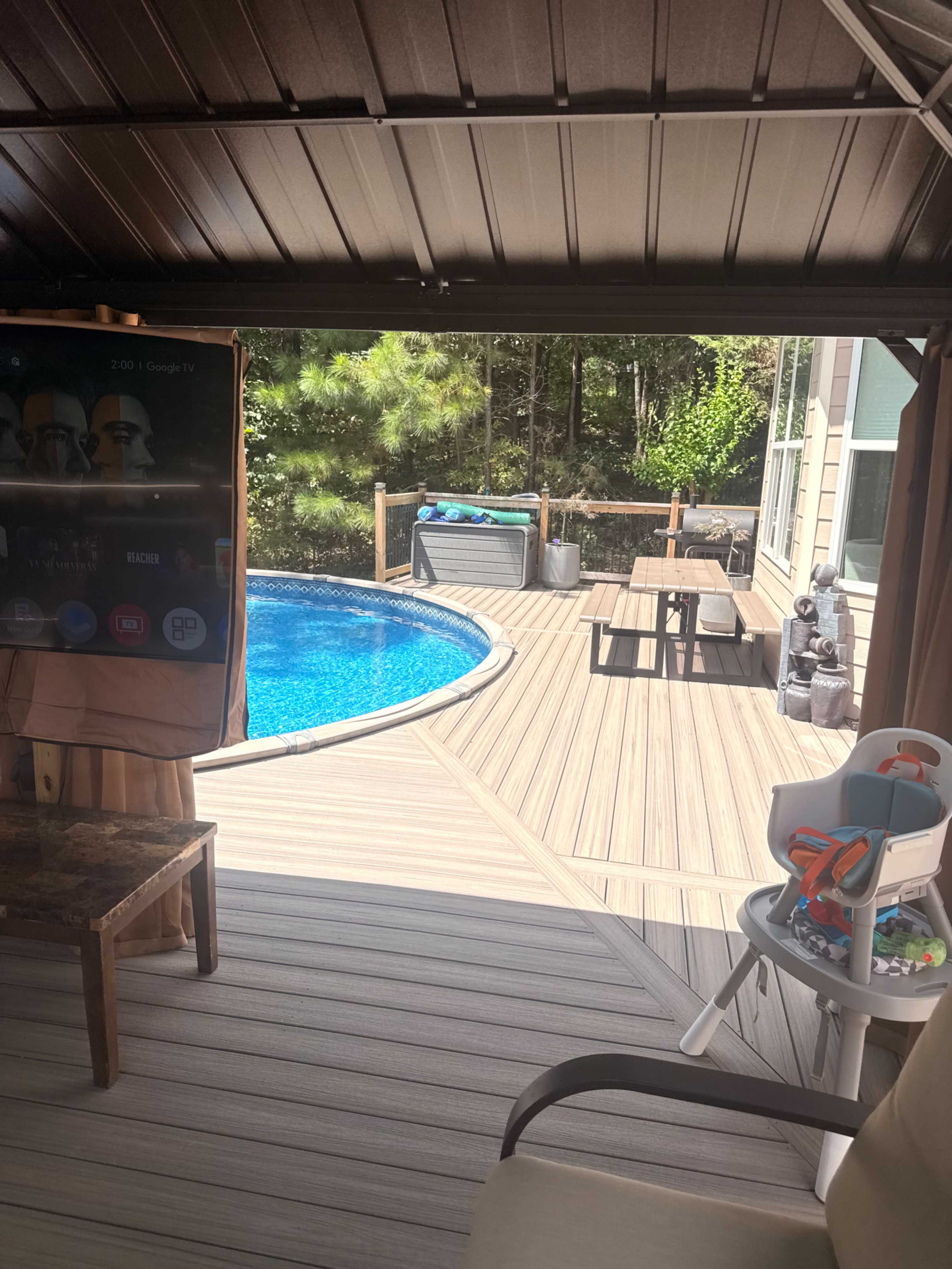 The image shows a backyard patio featuring a swimming pool, outdoor furniture, and a television mounted on a nearby structure.
