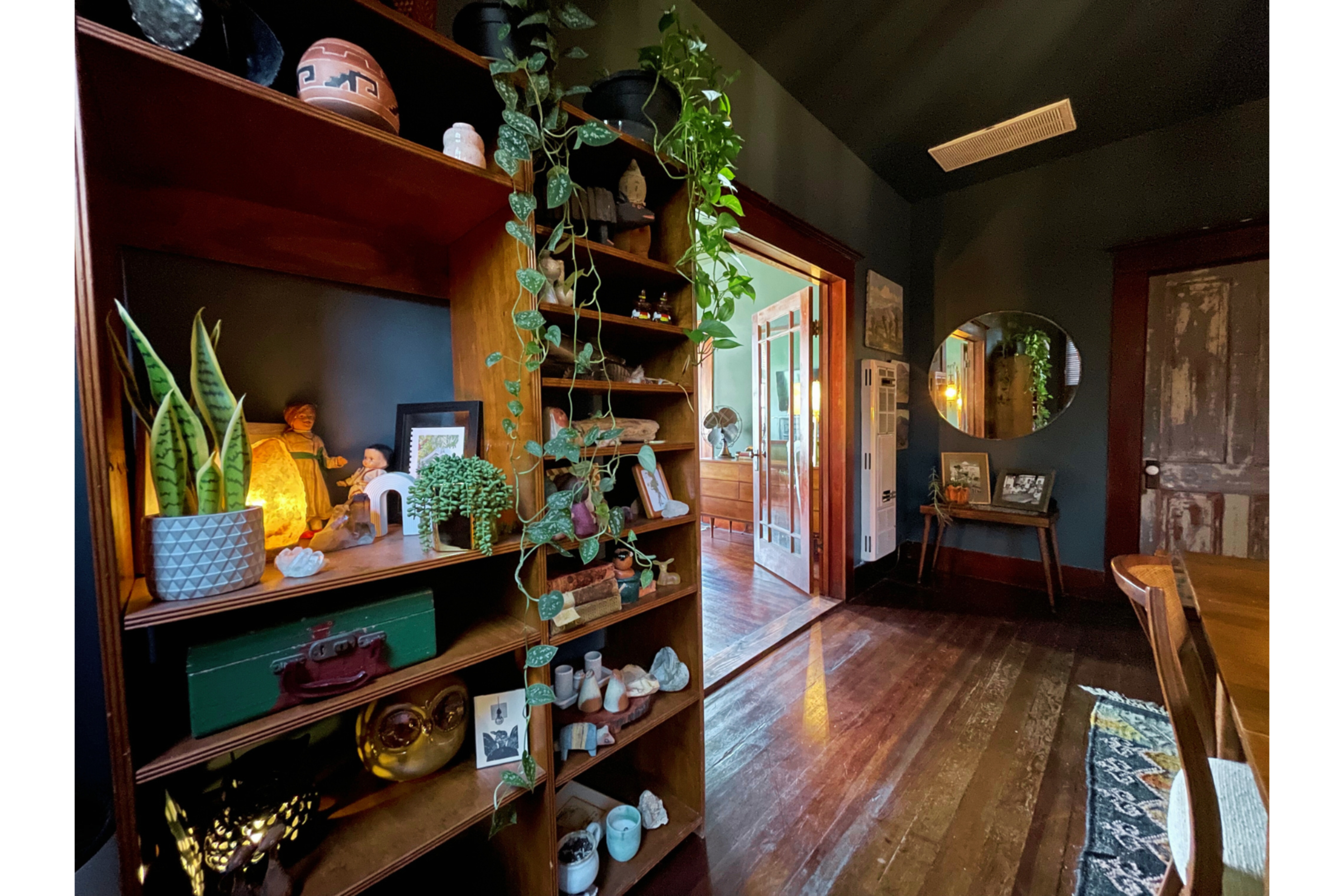 A wooden bookshelf filled with decorative items and plants stands in a room with dark walls, leading to a doorway that opens into a brighter space.
