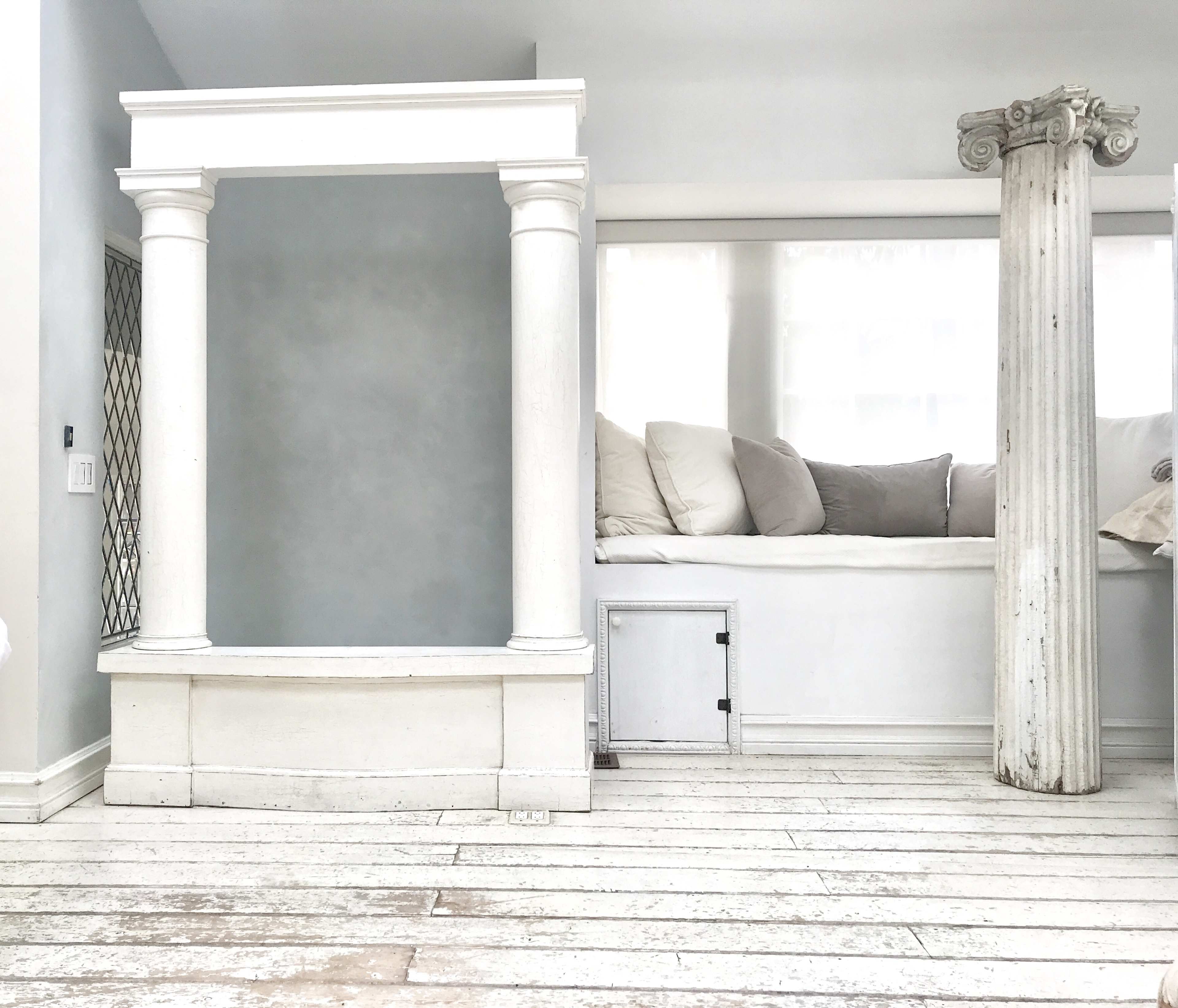 A minimalist interior space featuring a white frame resembling a doorway and a decorative column beside a cushioned area along the wall.