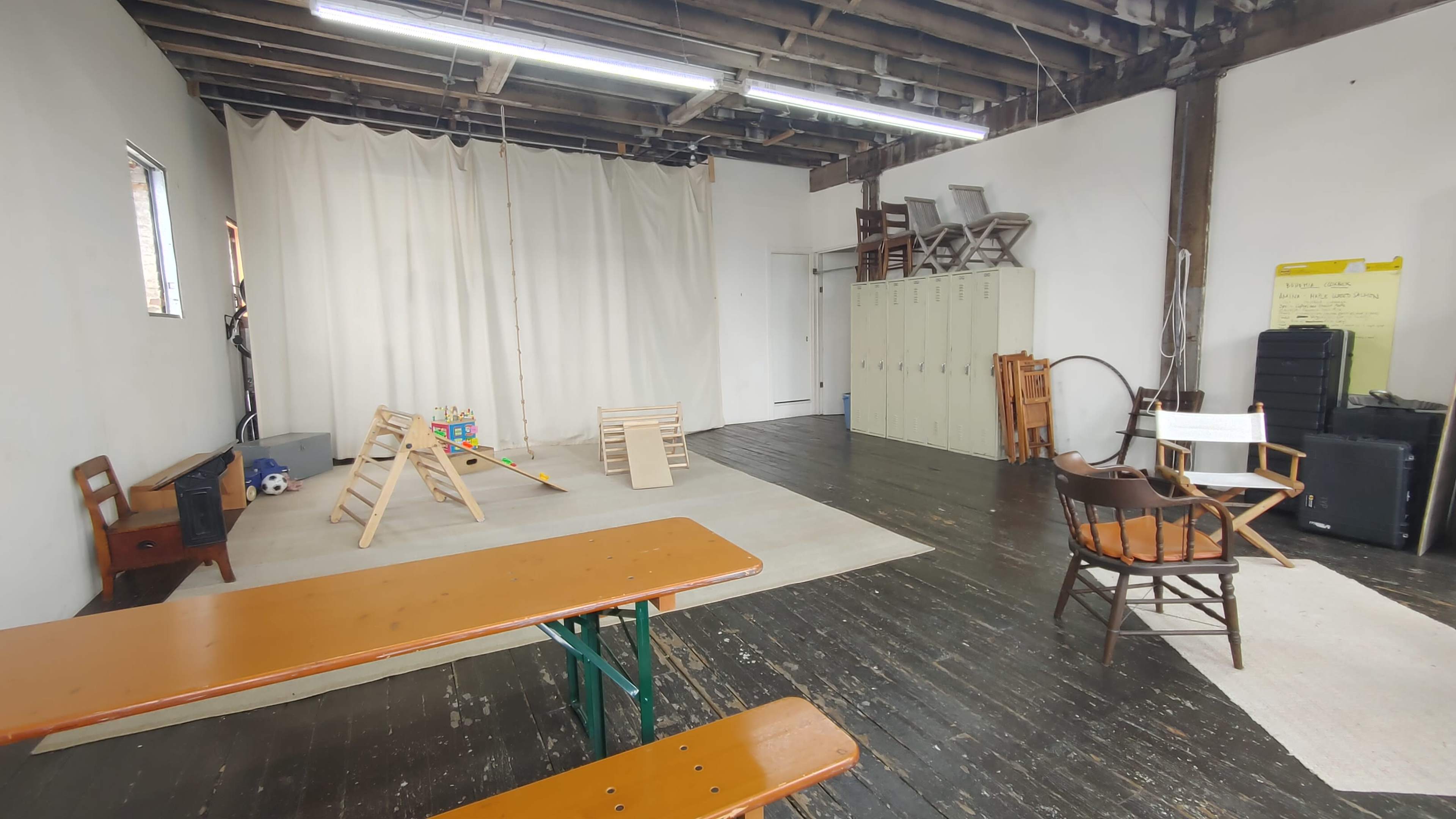 The image shows an empty room with wooden floors, a few play structures, a white curtain backdrop, and some furniture including wooden tables and chairs.