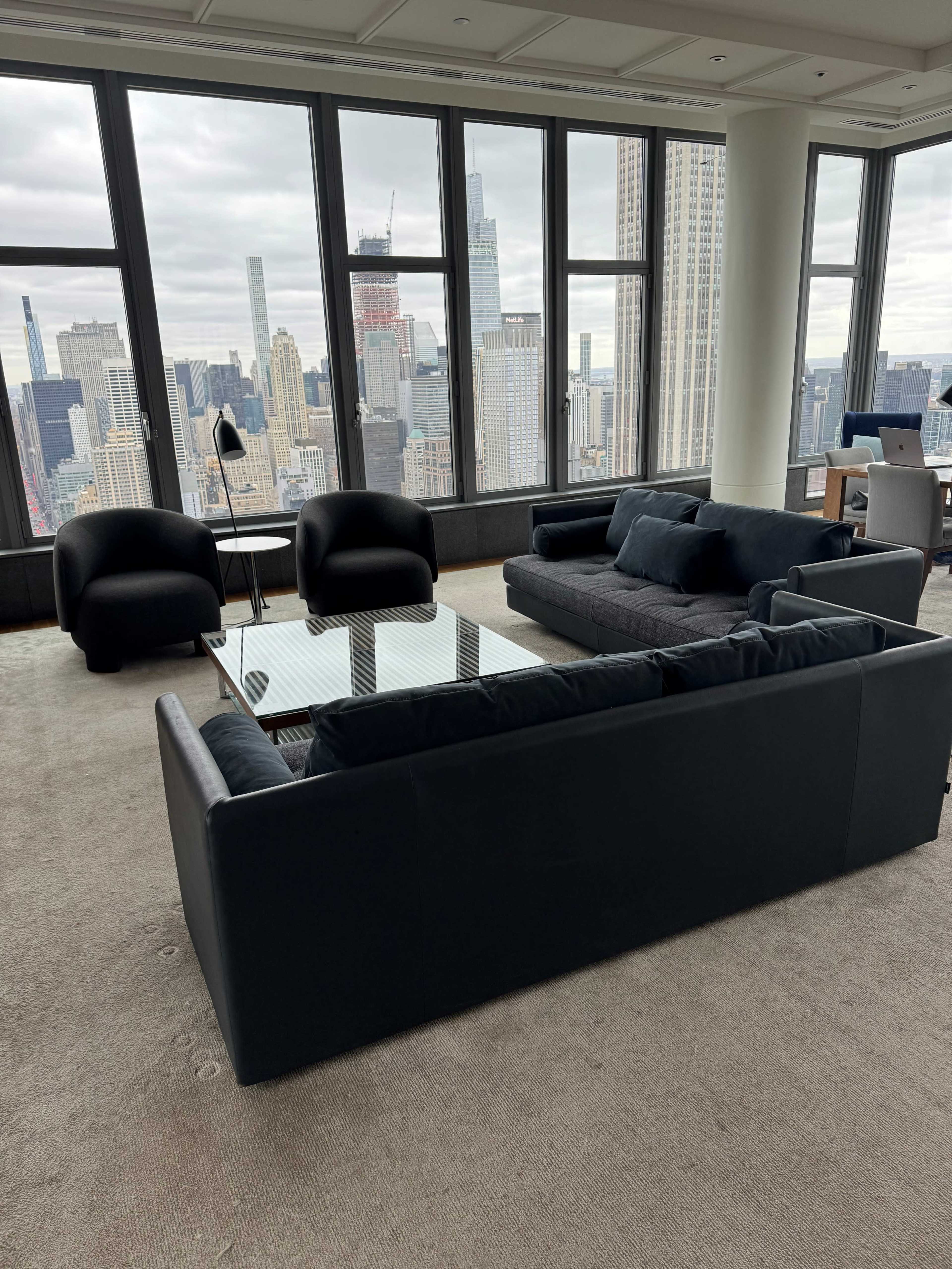 A modern living area features a large black sectional sofa, two armchairs, and a glass coffee table, all set against a backdrop of expansive city skyline views through large windows.