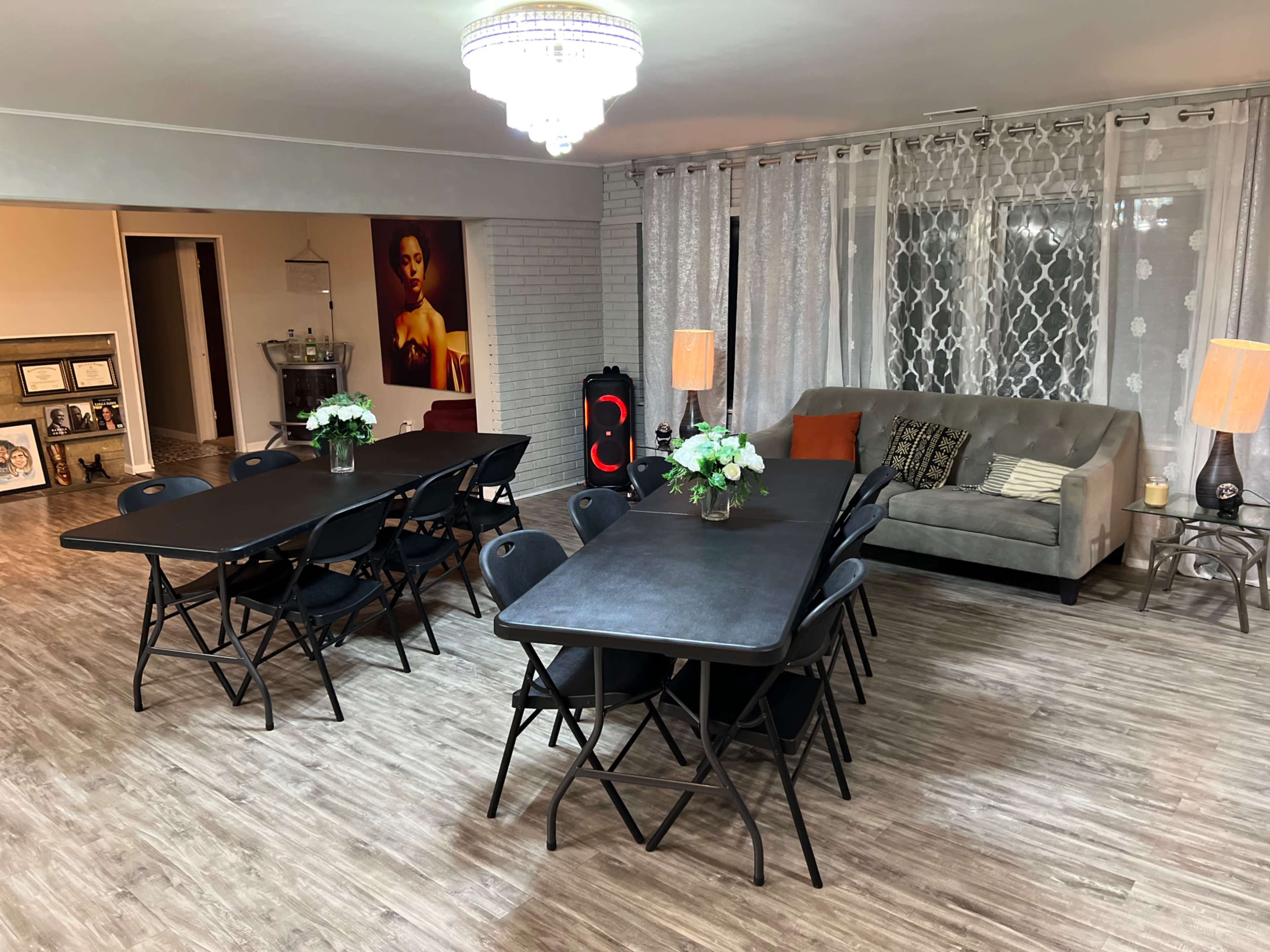 A spacious room featuring two long tables surrounded by folding chairs, a comfortable couch, decorative lamps, and a chandelier overhead.