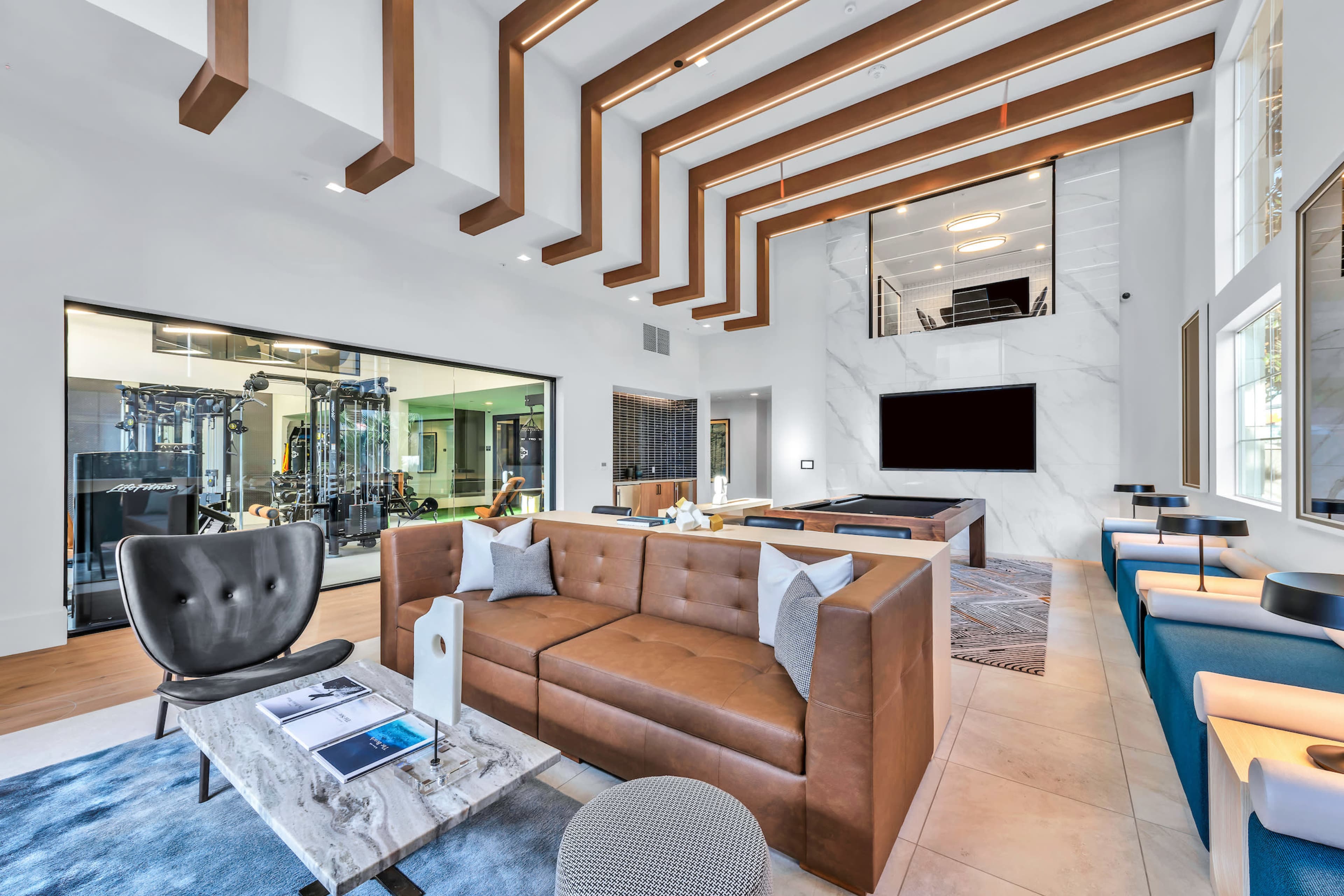 A modern lounge area featuring a brown leather sectional sofa, a coffee table, and a large wall-mounted TV, with a view of a gym through a glass wall.