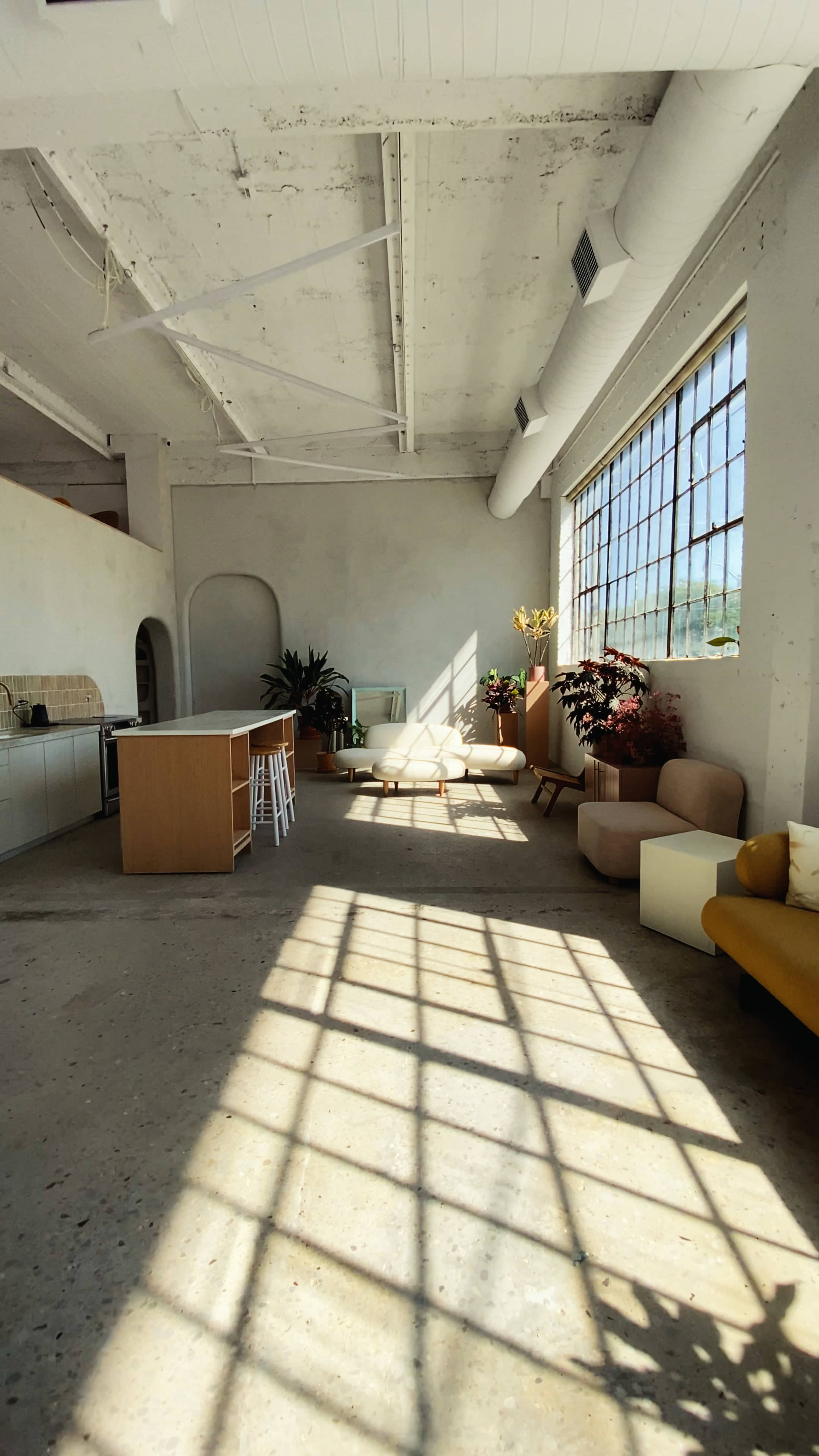 A spacious room features large windows casting shadows on the concrete floor, with various seating areas and plants arranged throughout.