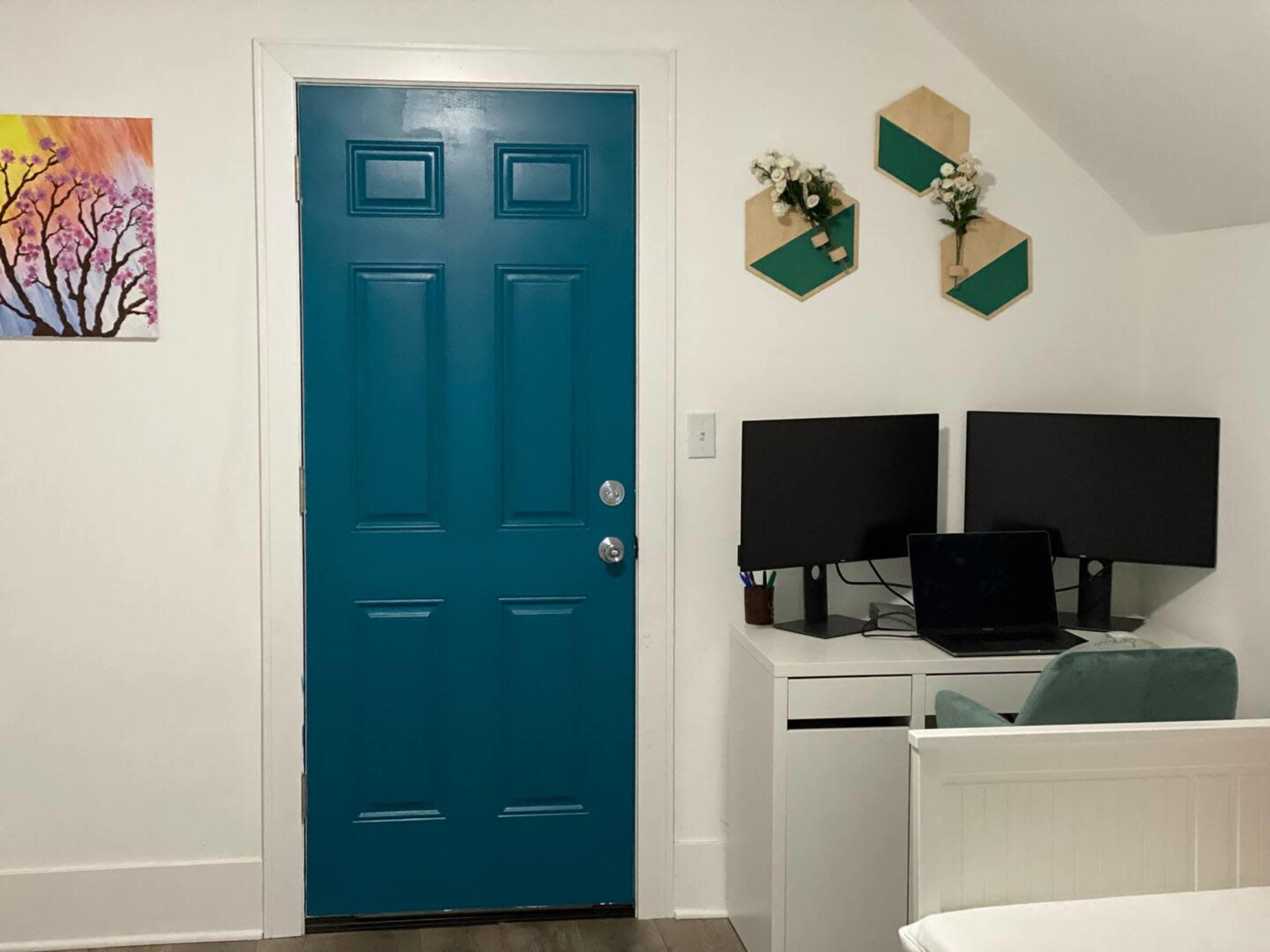 A teal door is positioned against a white wall, next to a desk with two monitors and a laptop, while decorative hexagonal shelves with plants are mounted above.