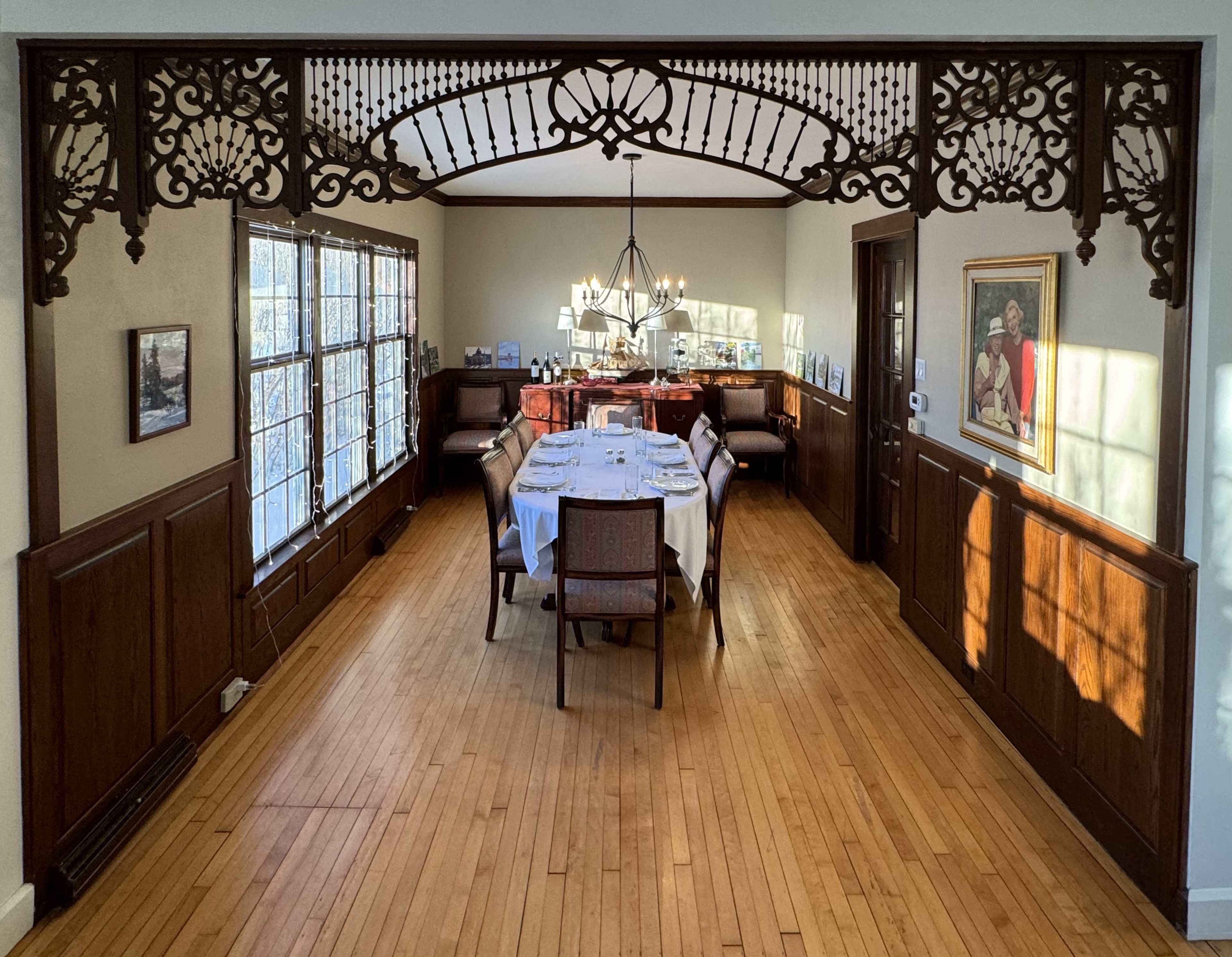 A dining room features a long table set for a meal, surrounded by chairs, with large windows letting in natural light and intricate woodwork framing the entrance.