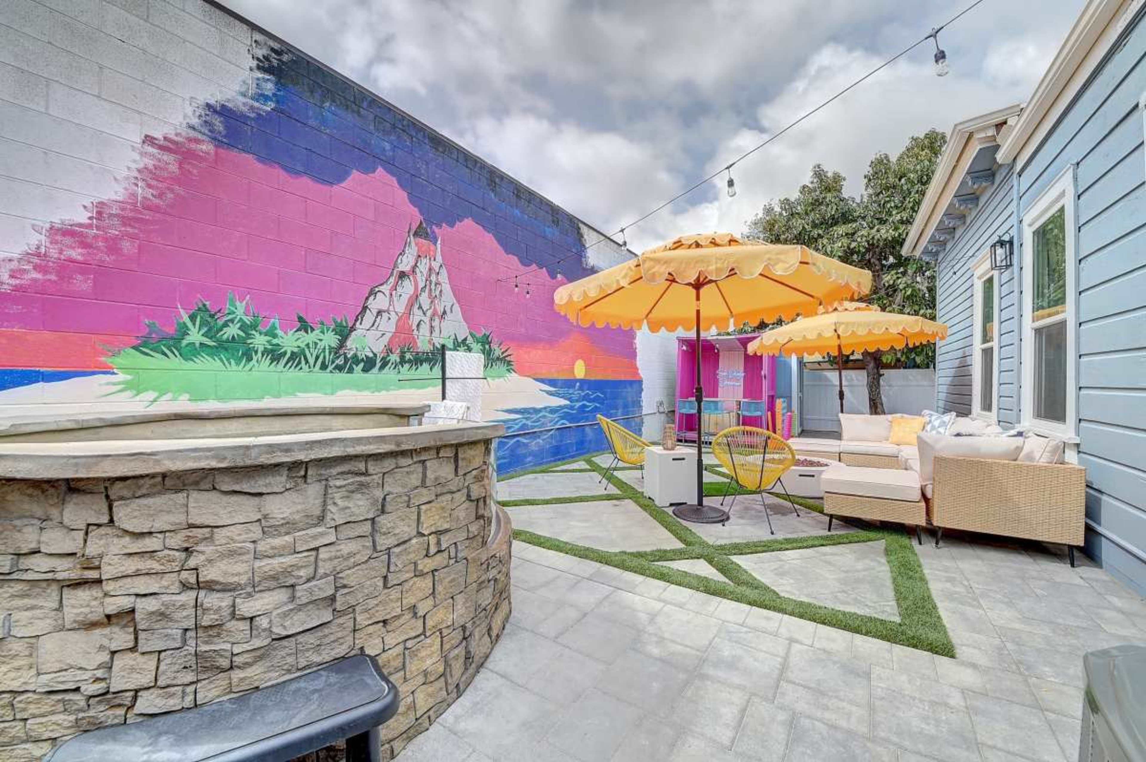 The outdoor space features a stone wall bar, vibrant mural, yellow umbrellas over seating, and a well-maintained lawn area.