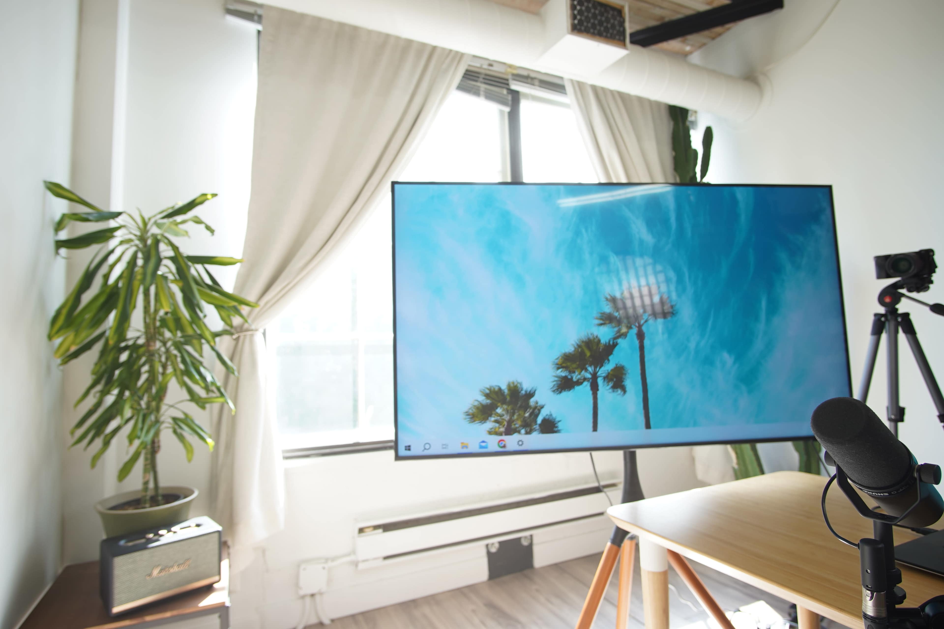 A bright room features a large monitor displaying a scene of palm trees, with a microphone and a camera set up on a table nearby.