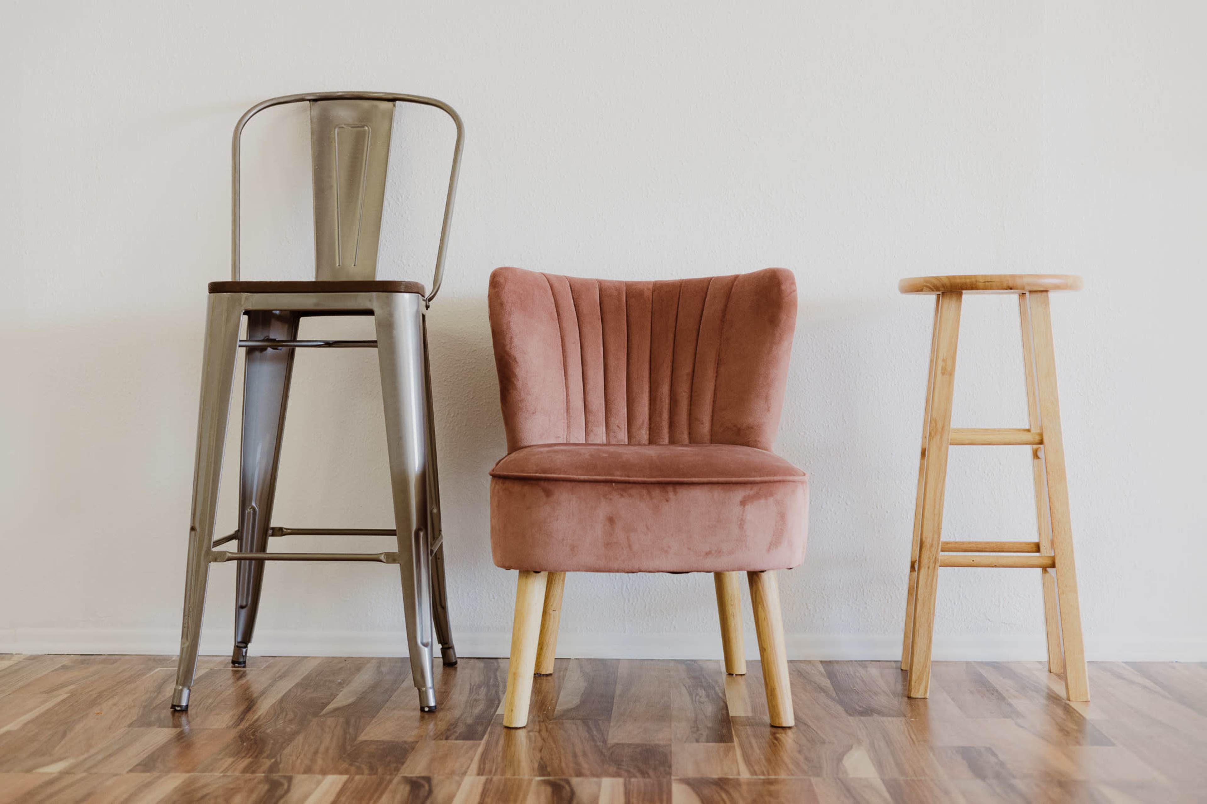 Three distinct chairs: a metal bar stool, a plush pink armchair, and a wooden stool, arranged side by side against a blank wall.