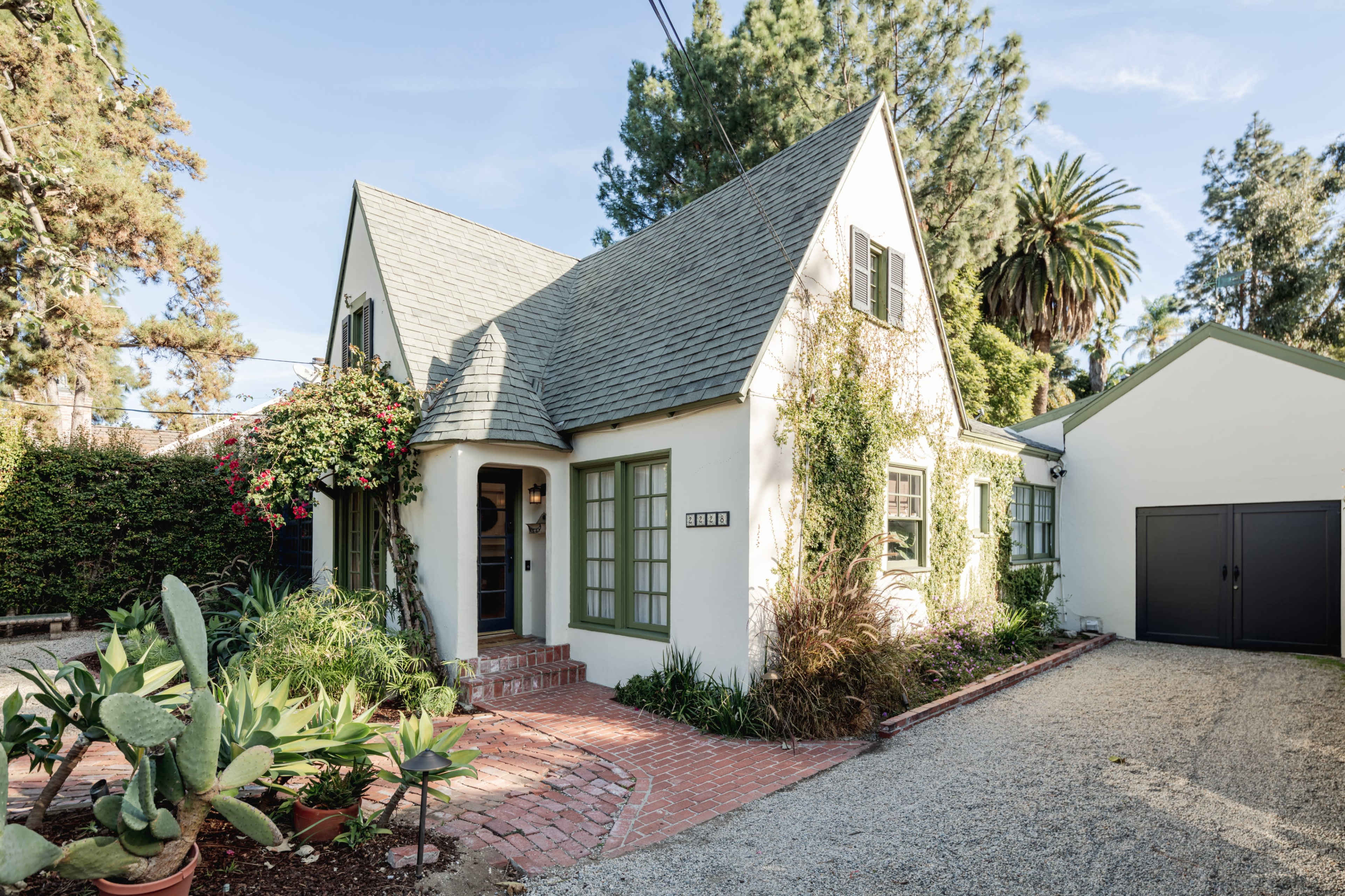 A single-story house with a steep pitched roof, surrounded by greenery and featuring a brick walkway and gravel driveway.
