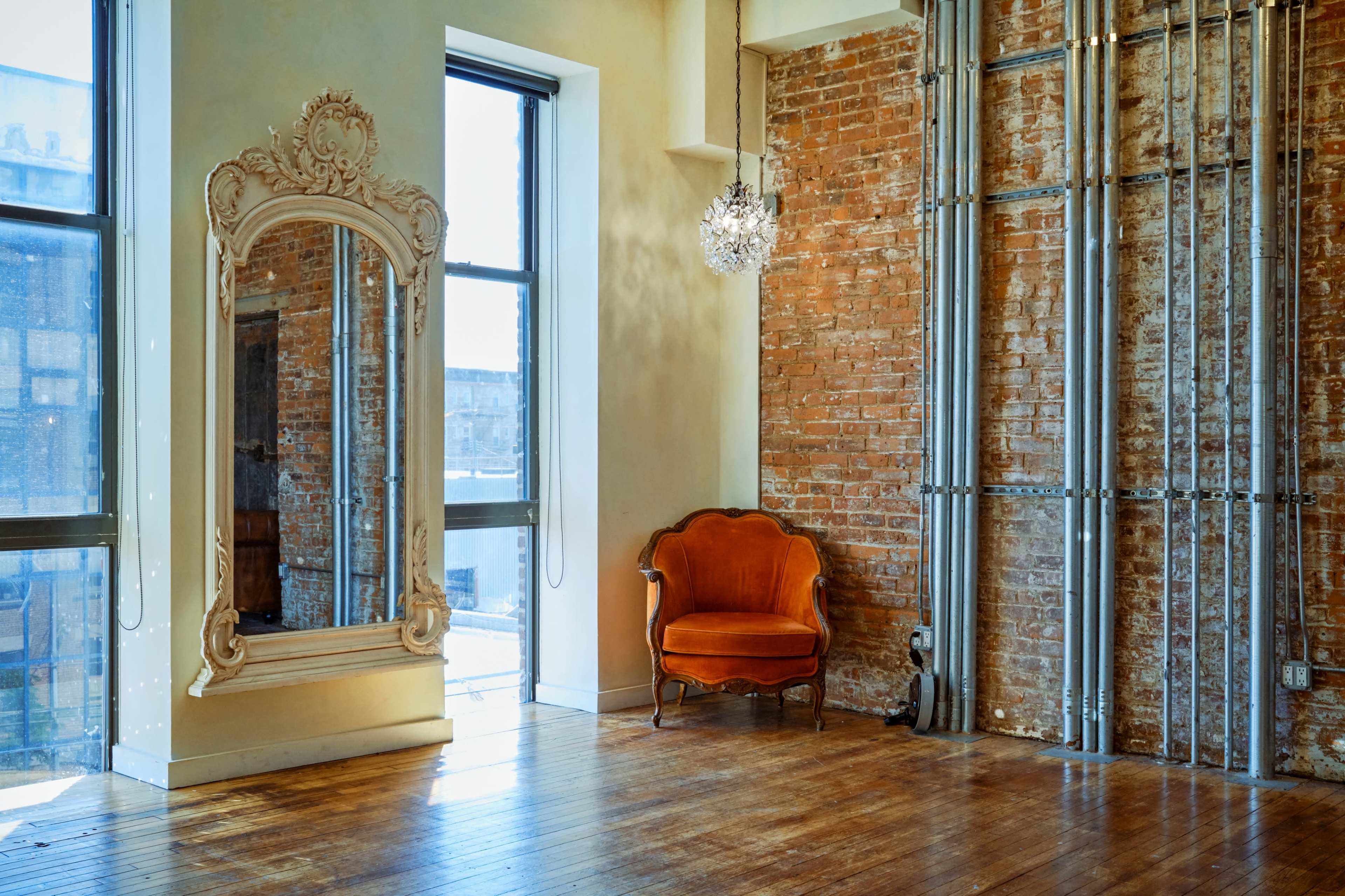 The image shows a spacious room with exposed brick walls, a large decorative mirror, a vintage orange chair, and industrial piping.