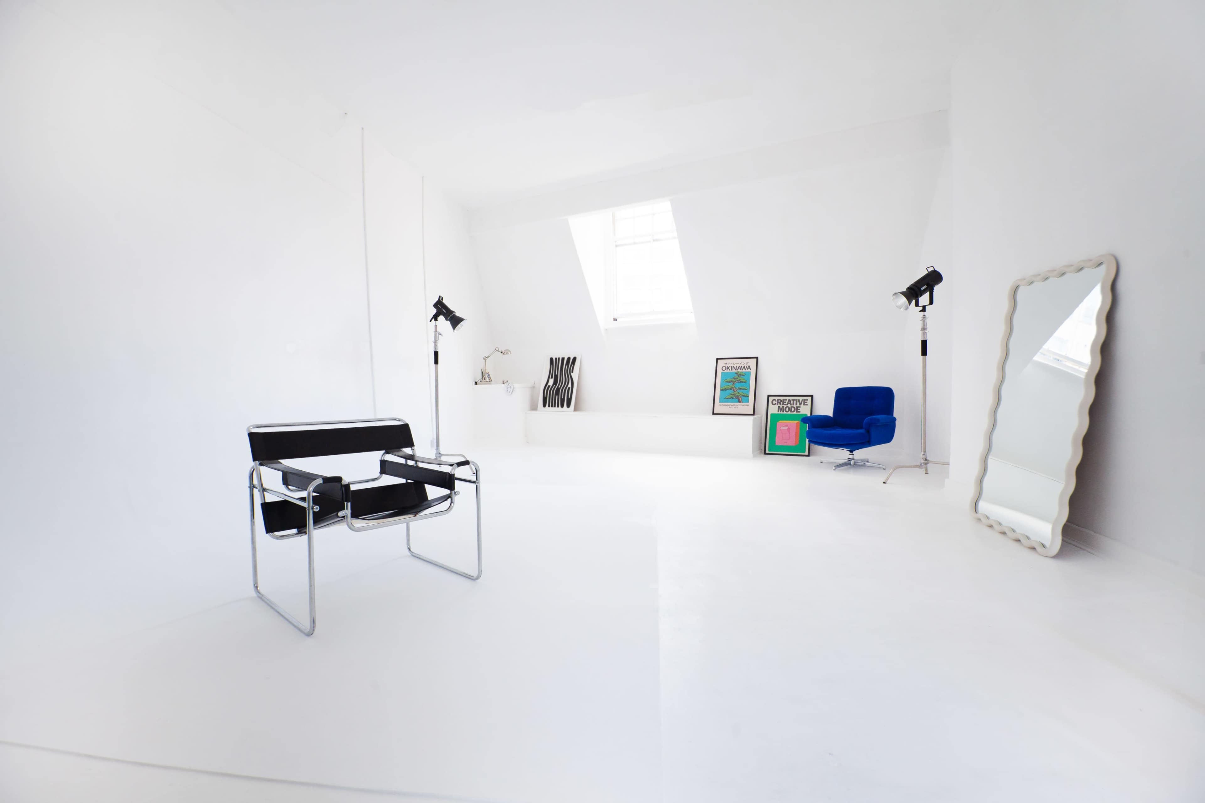 The image shows a sparsely furnished, bright white room featuring a black and chrome chair, a blue armchair, two studio lights, and a large mirror.