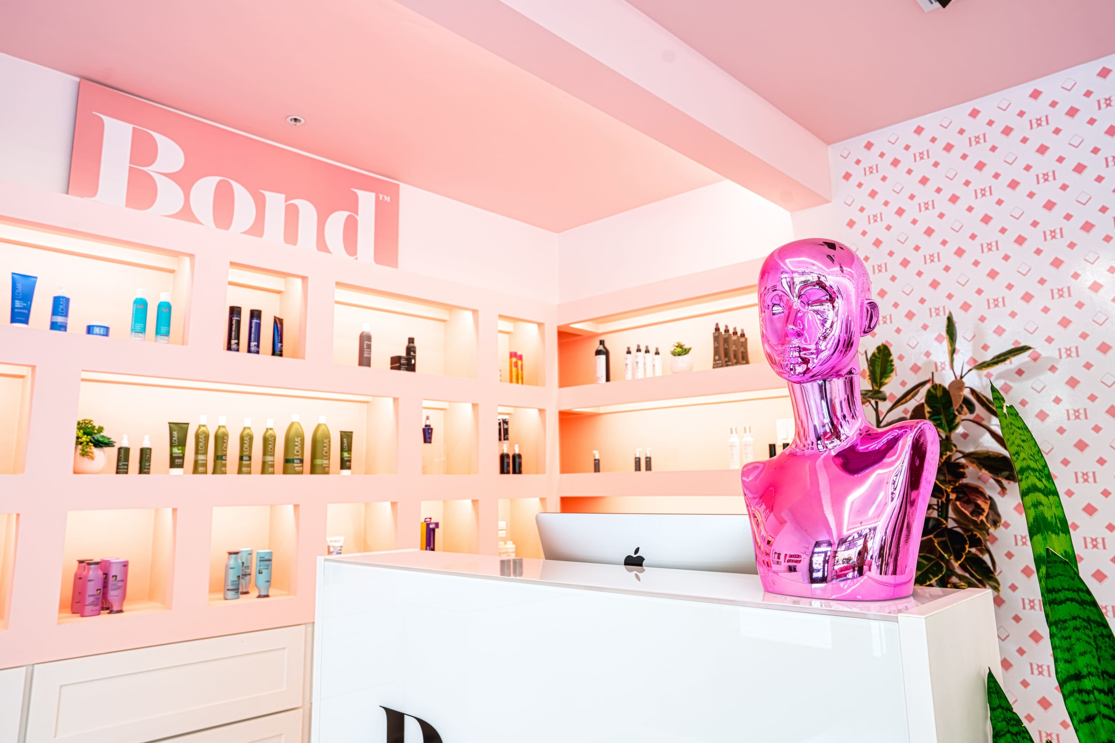 A stylish beauty salon interior featuring a pink reception desk with a transparent pink mannequin head and neatly arranged shelving displaying various beauty products against a pink and white patterned wall.