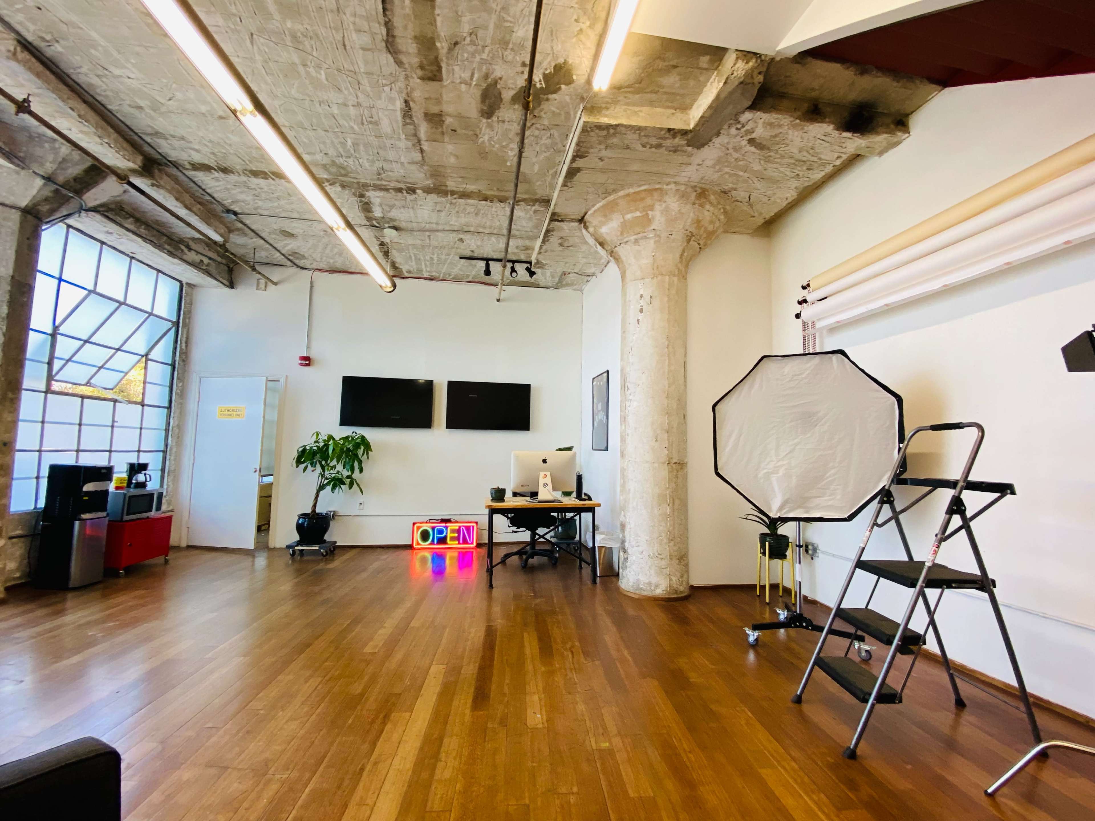 A spacious studio featuring hardwood floors, exposed concrete pillars, a desk with computer equipment, and a large softbox on a stand.
