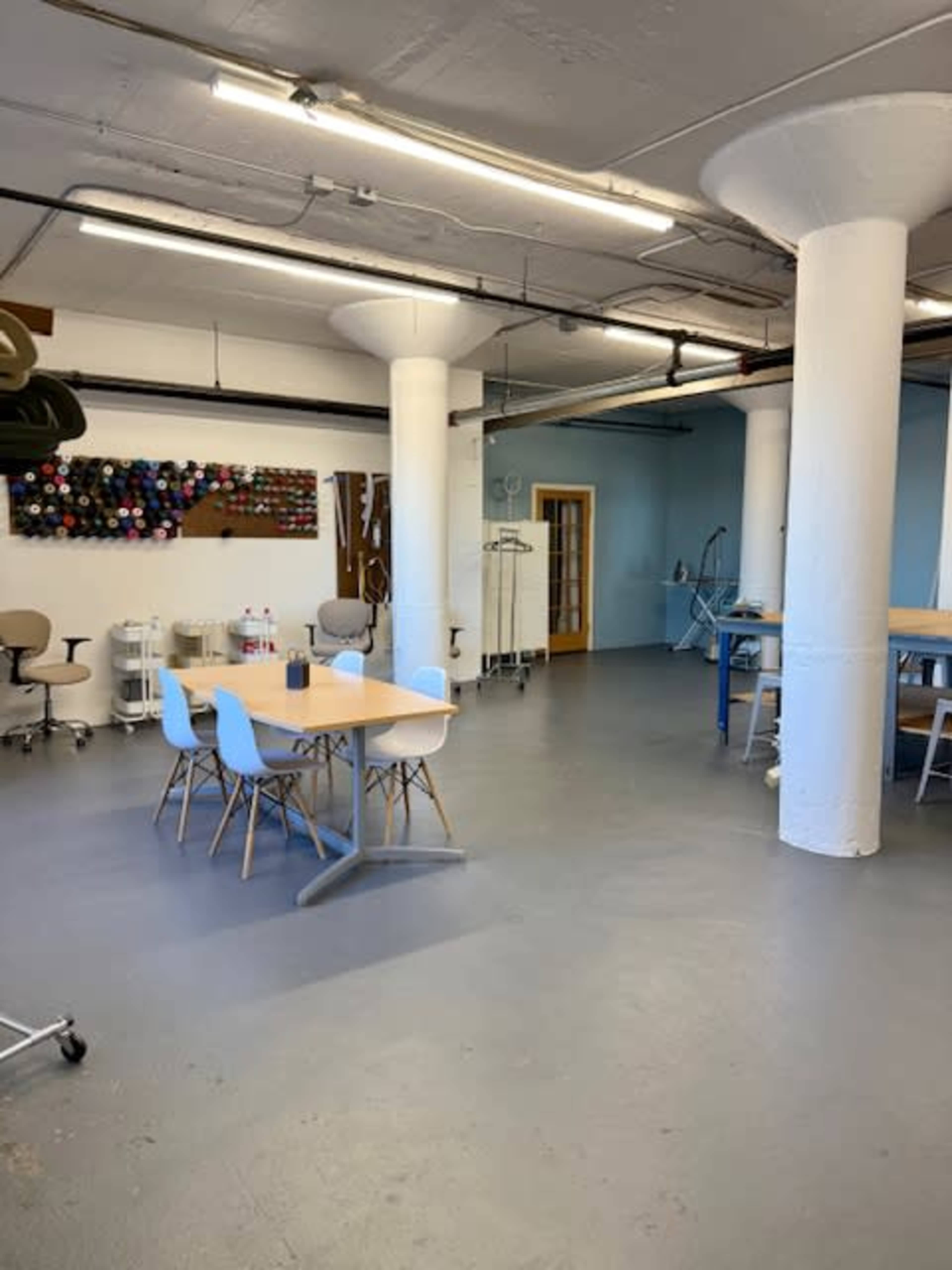 The image shows a spacious workshop or studio area with several tables, white walls, and large supporting columns.