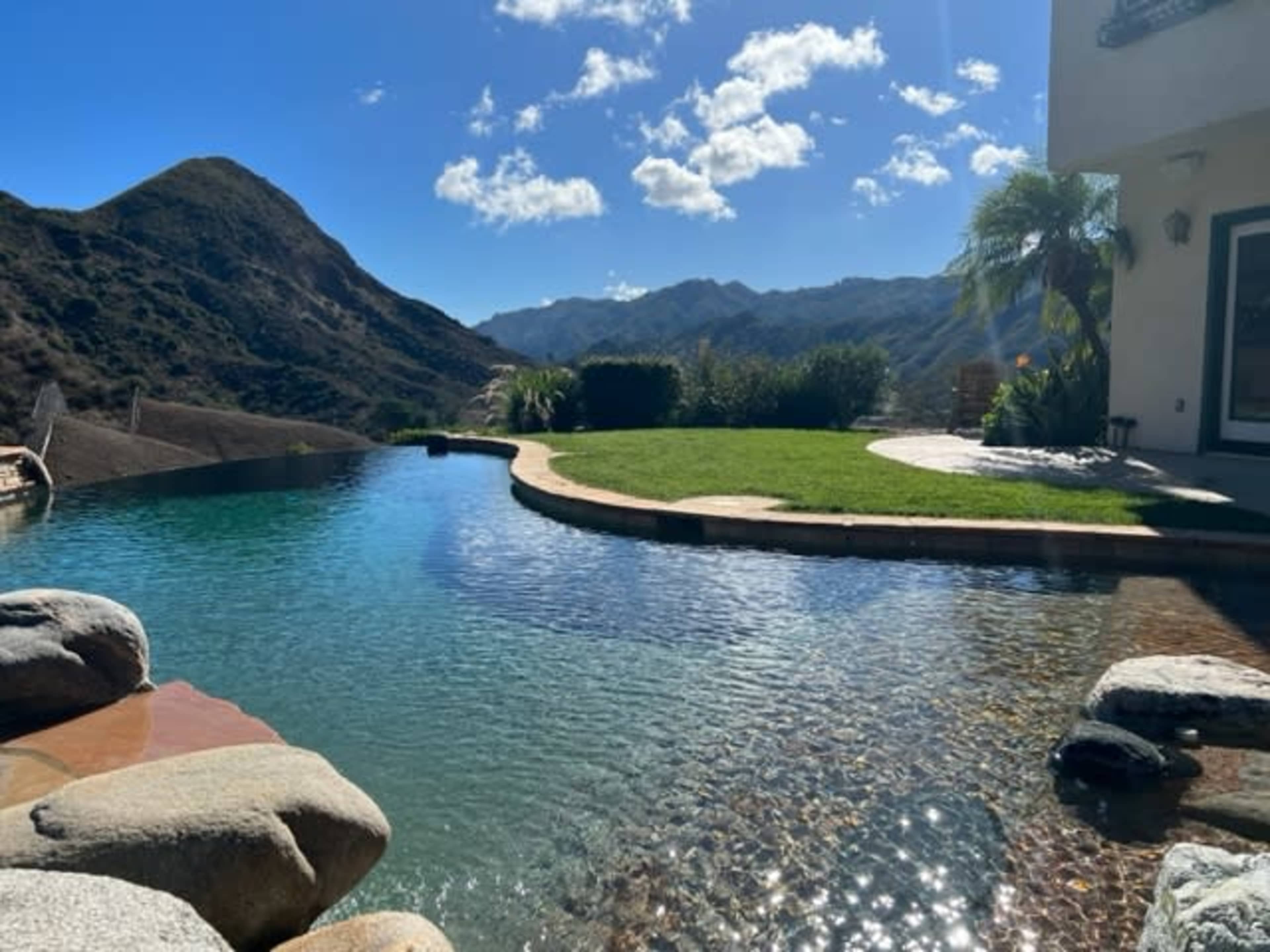 A pool with clear water beside a landscaped lawn and mountains in the background under a partly cloudy sky.