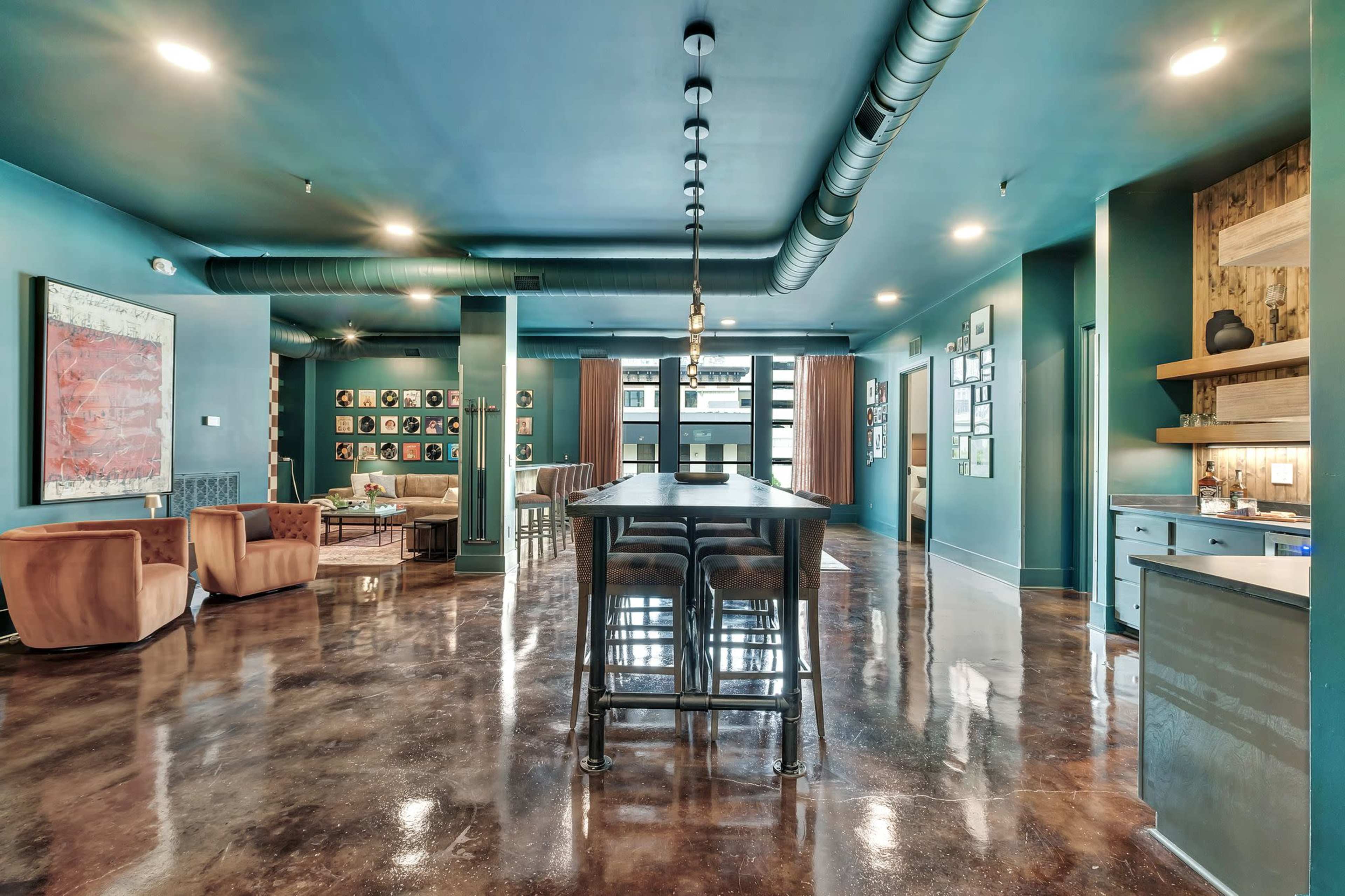 The image shows a modern lounge area with a long bar table, stylish seating, and a polished concrete floor, illuminated by overhead lighting.