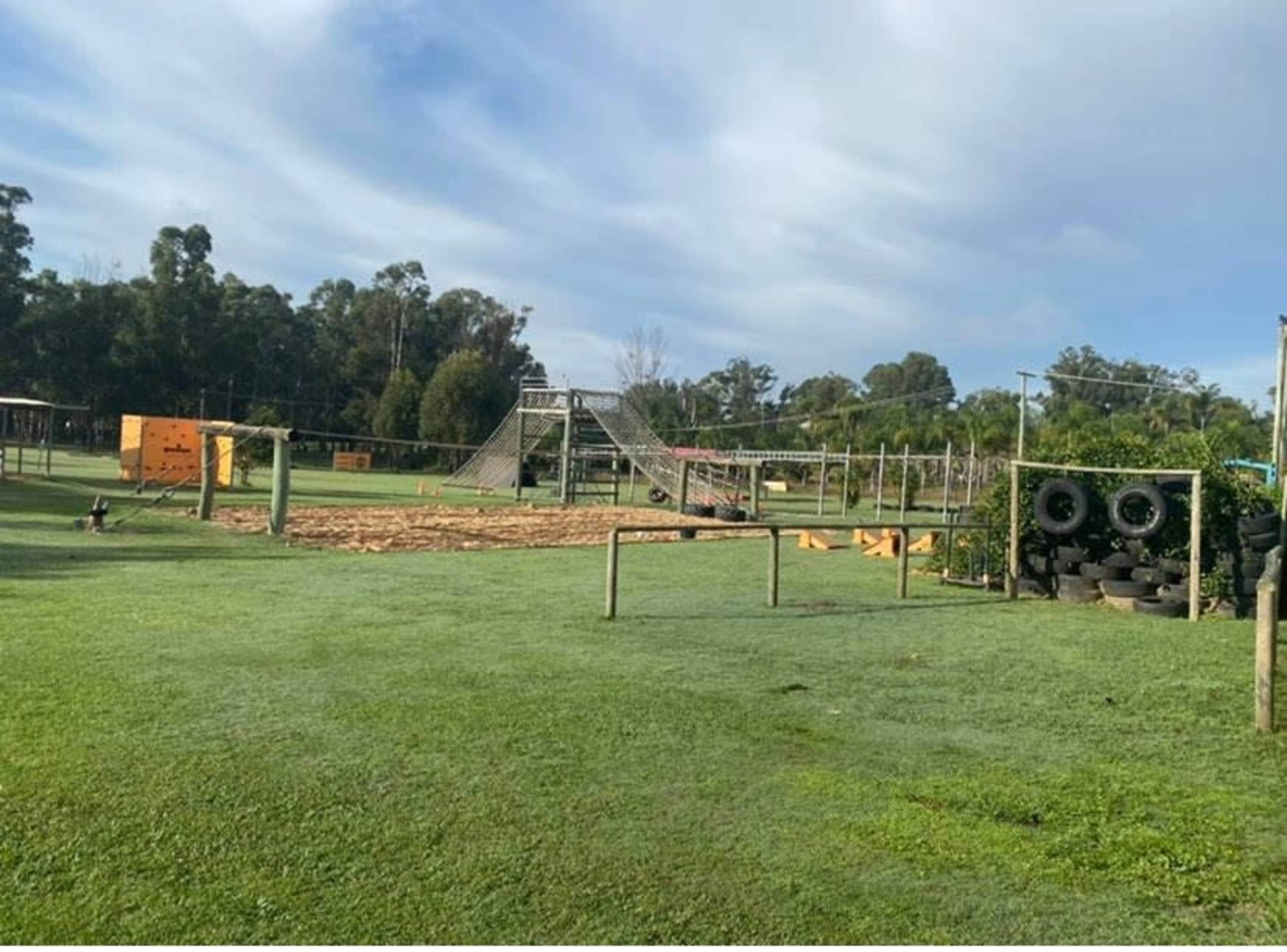 A playground with various climbing structures and equipment set on a grassy field, surrounded by trees.