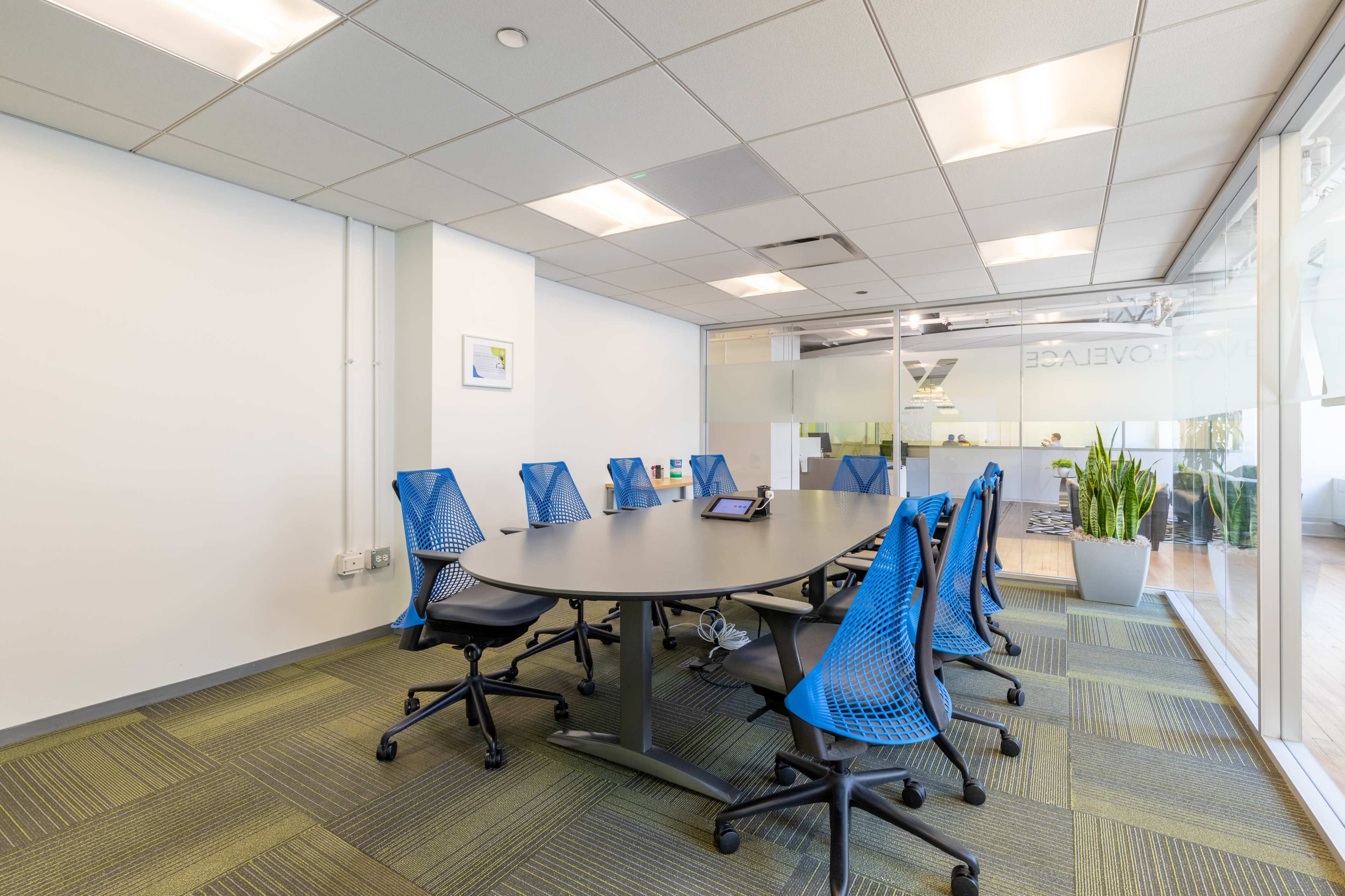 The image shows a modern conference room with a circular table surrounded by eight blue mesh office chairs and a large window letting in natural light.