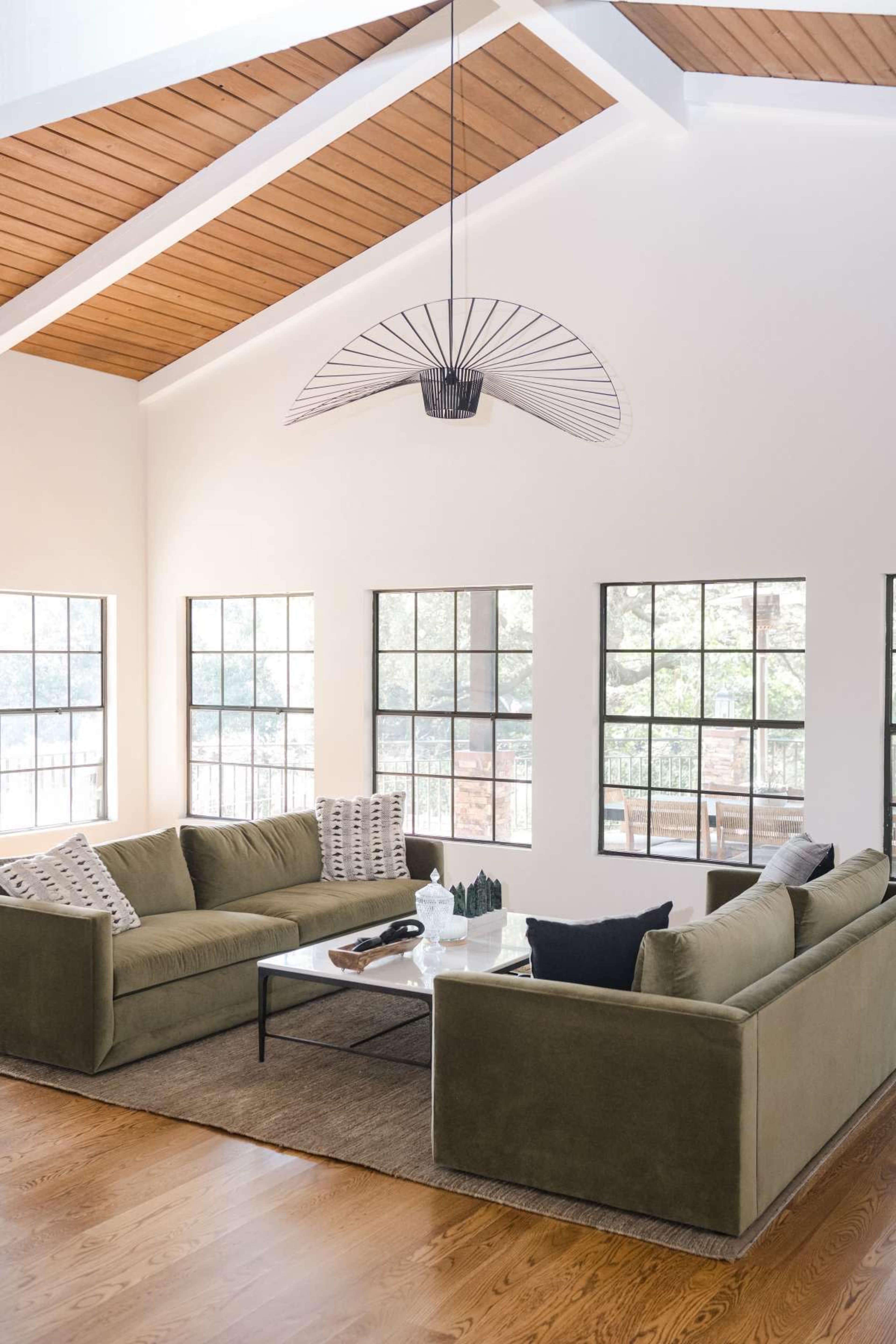 A spacious living room features two olive-green sofas arranged around a coffee table, with large windows allowing natural light and a sloped wooden ceiling above.