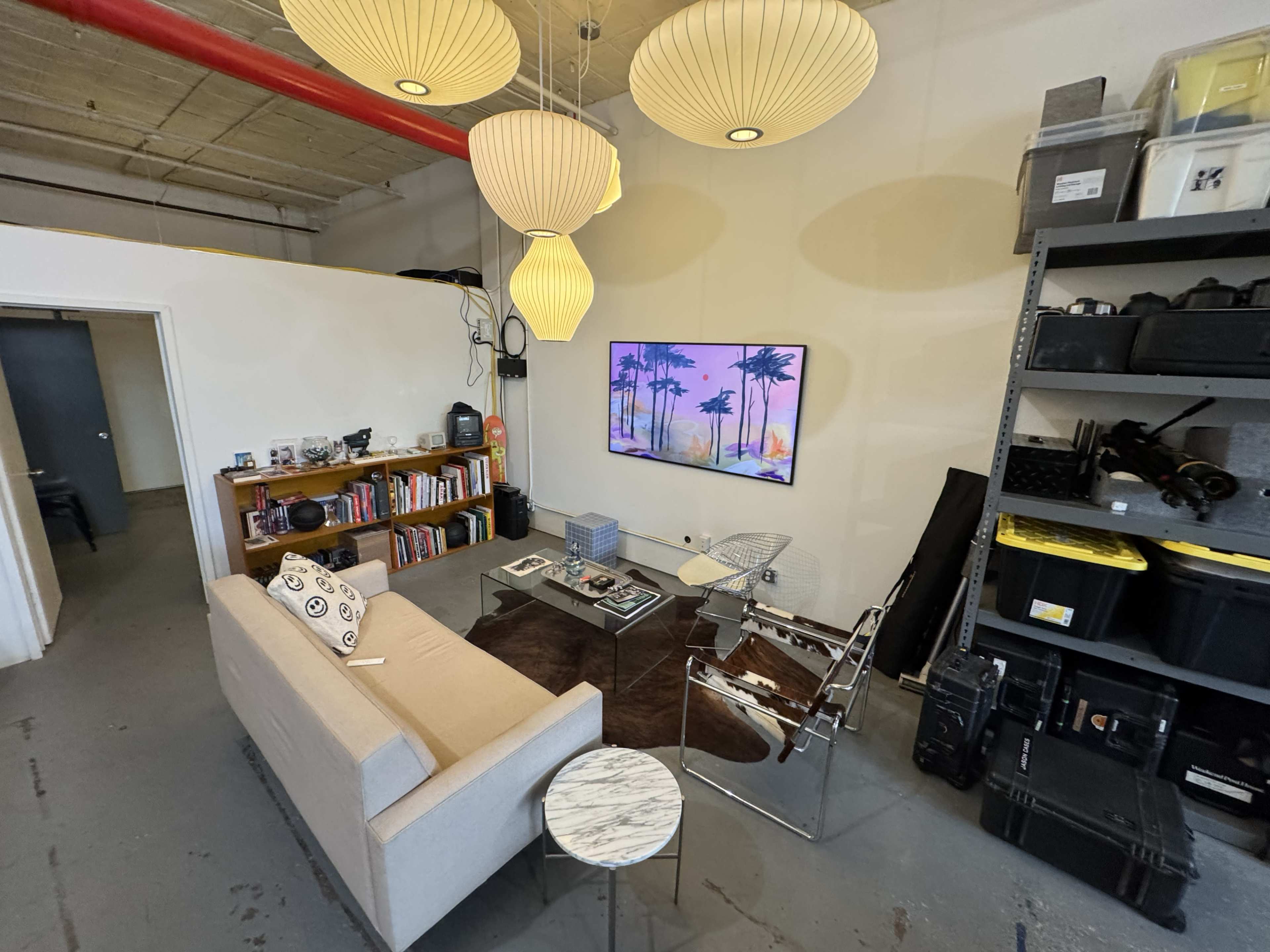 The image shows a modern office space featuring a sofa, a glass table, a wall-mounted television displaying a landscape, and shelving units with various equipment.