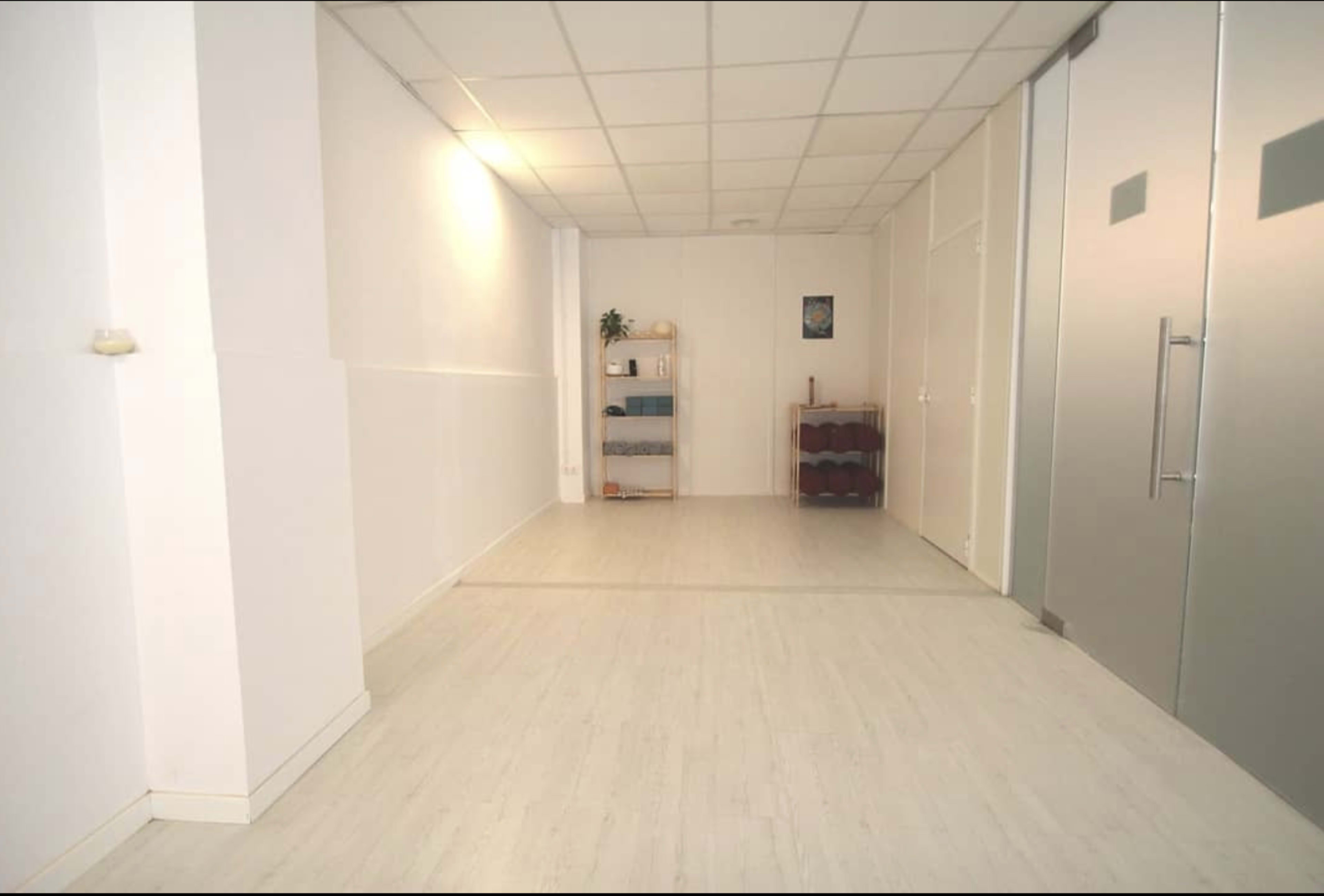 The image shows a spacious, well-lit room with white walls, a light-colored floor, and a shelf containing items on the left side.